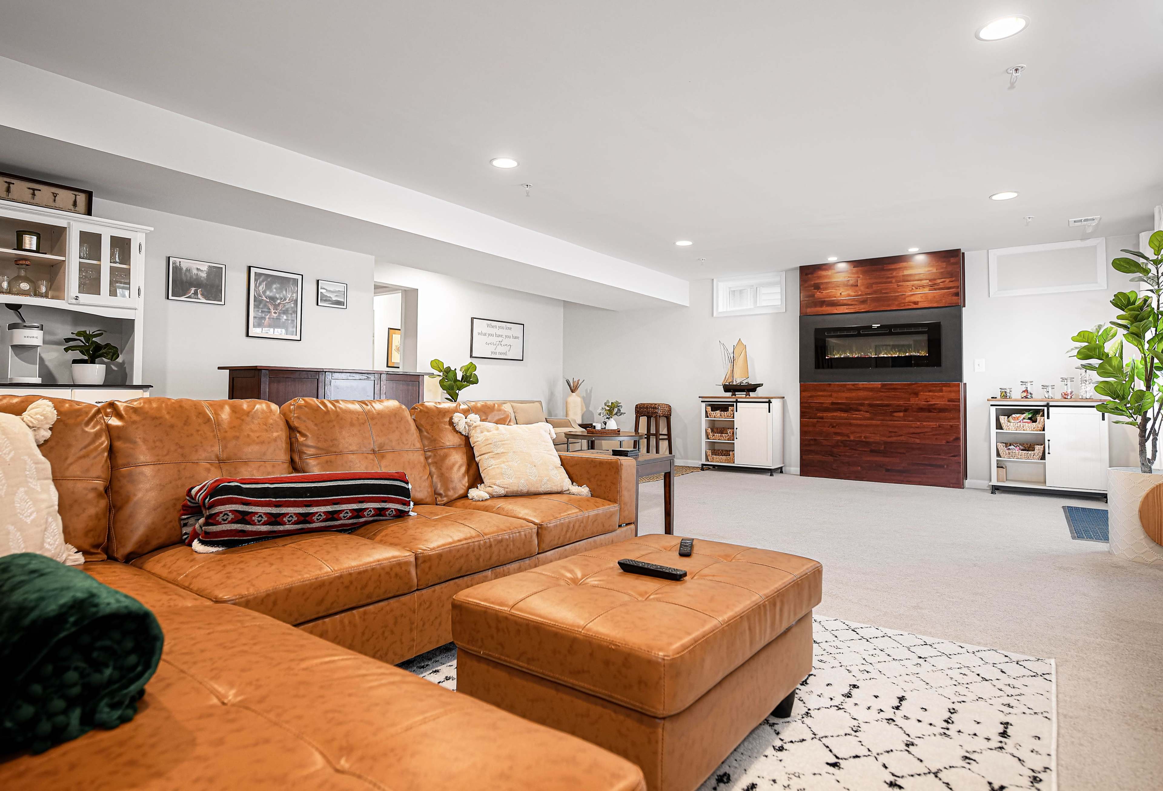 The image shows a spacious, modern basement living area with a brown leather sectional sofa, a fireplace, and a small bar area.