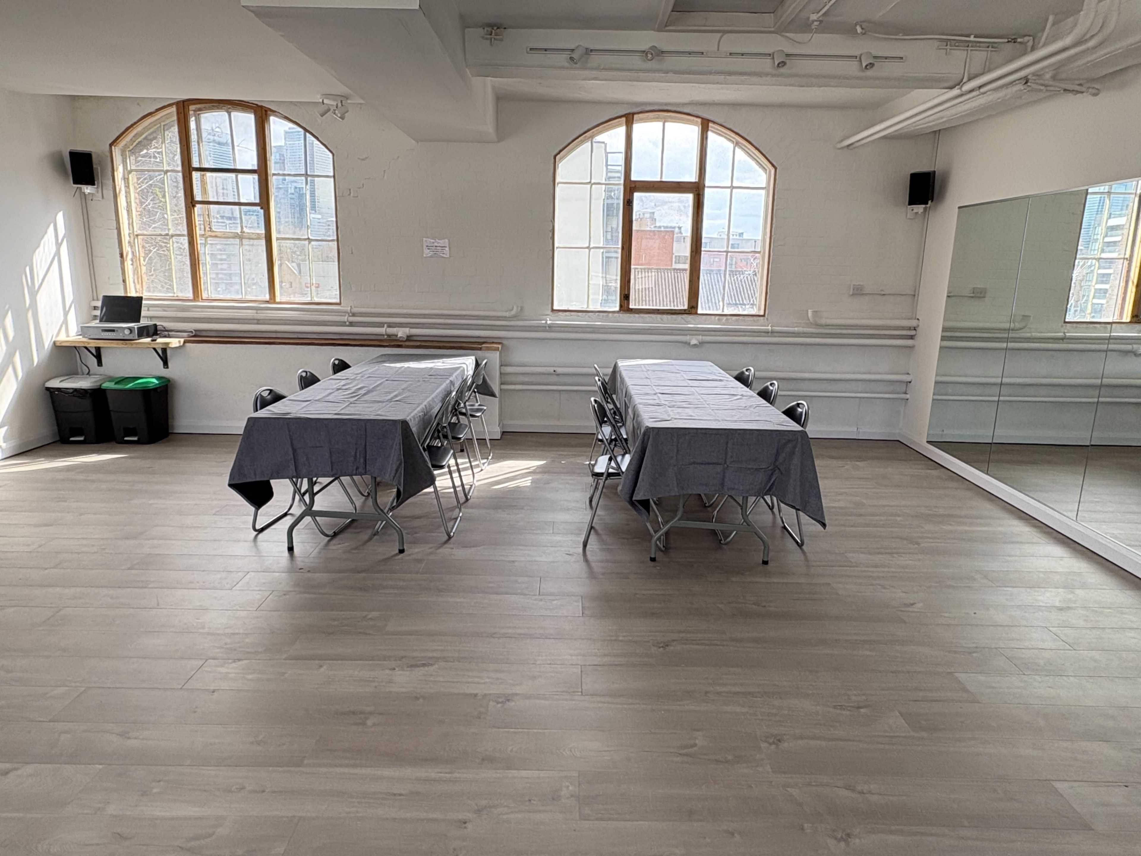The image shows a spacious room with two tables covered in gray tablecloths, surrounded by chairs, and large windows along one wall.