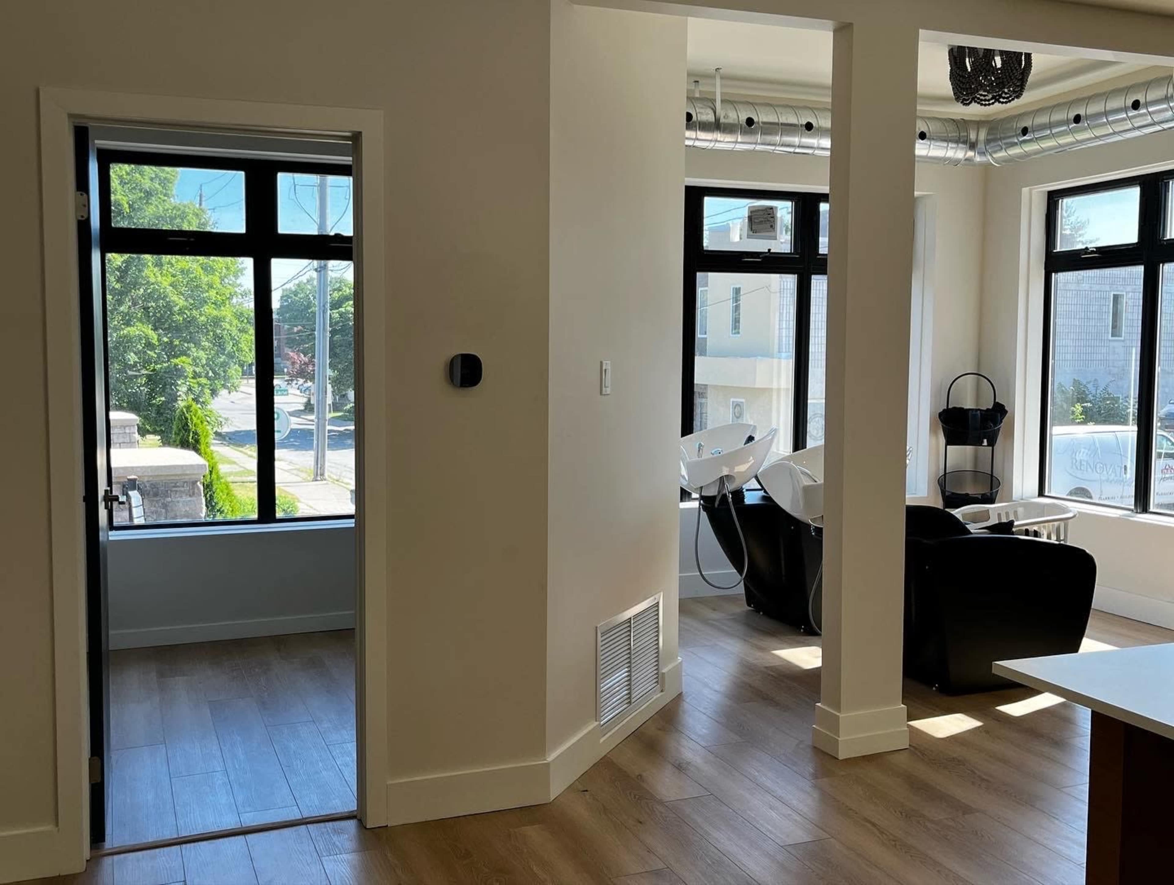 A modern interior space with a hallway leading to a room featuring two black salon chairs and large windows that provide natural light.