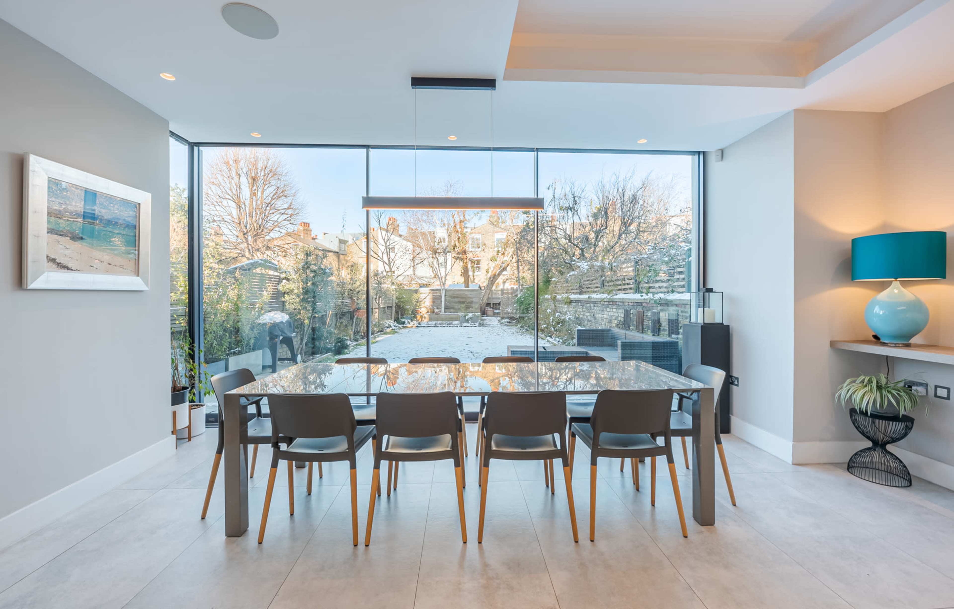 A modern dining room features a large glass table surrounded by six chairs, with a view of a garden through expansive windows.