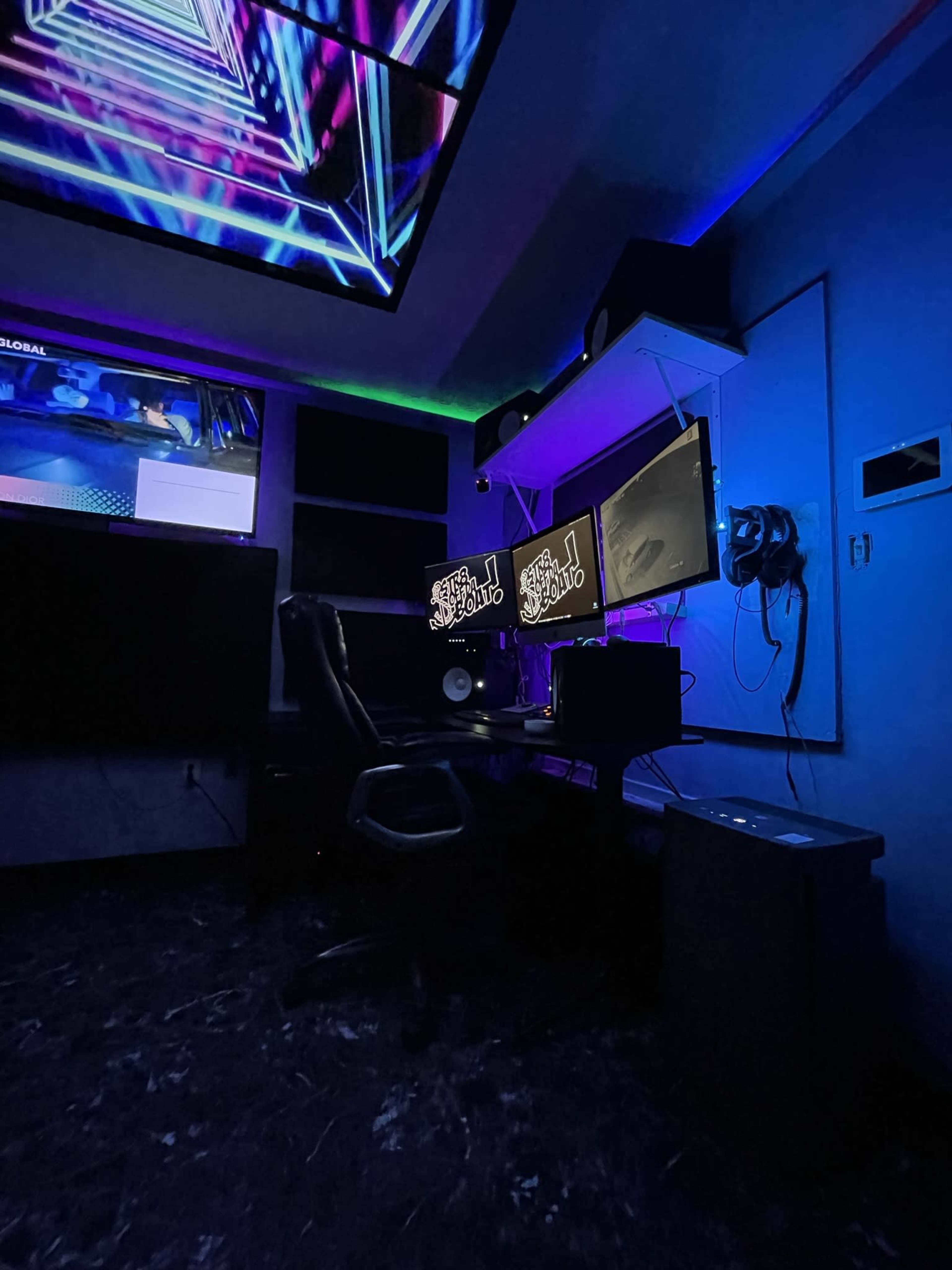 A dimly lit gaming or recording studio with multiple screens, a black chair, and colorful ambient lighting.