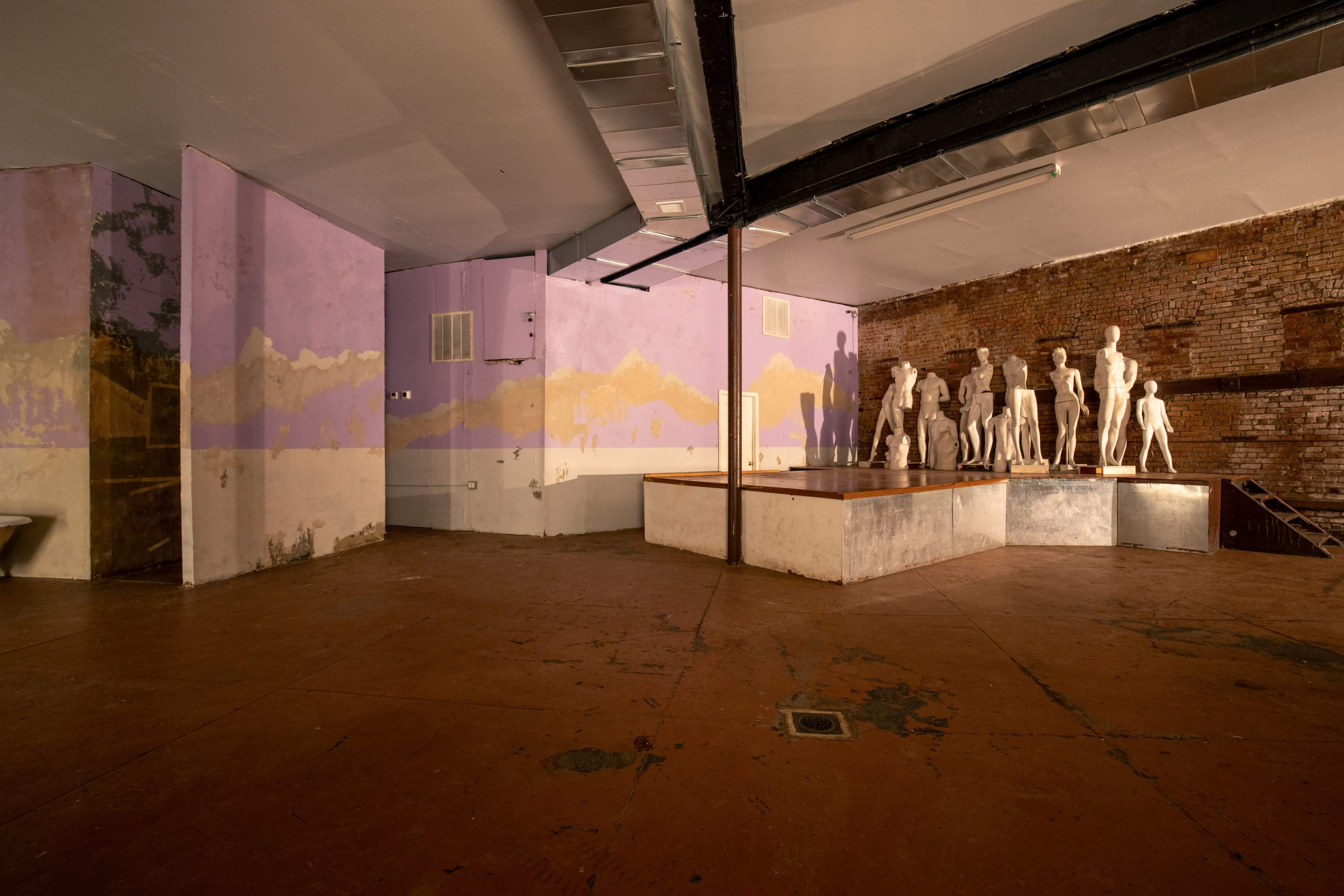 An empty room features a group of white mannequins on a raised wooden platform, with partially painted purple walls and exposed brick.