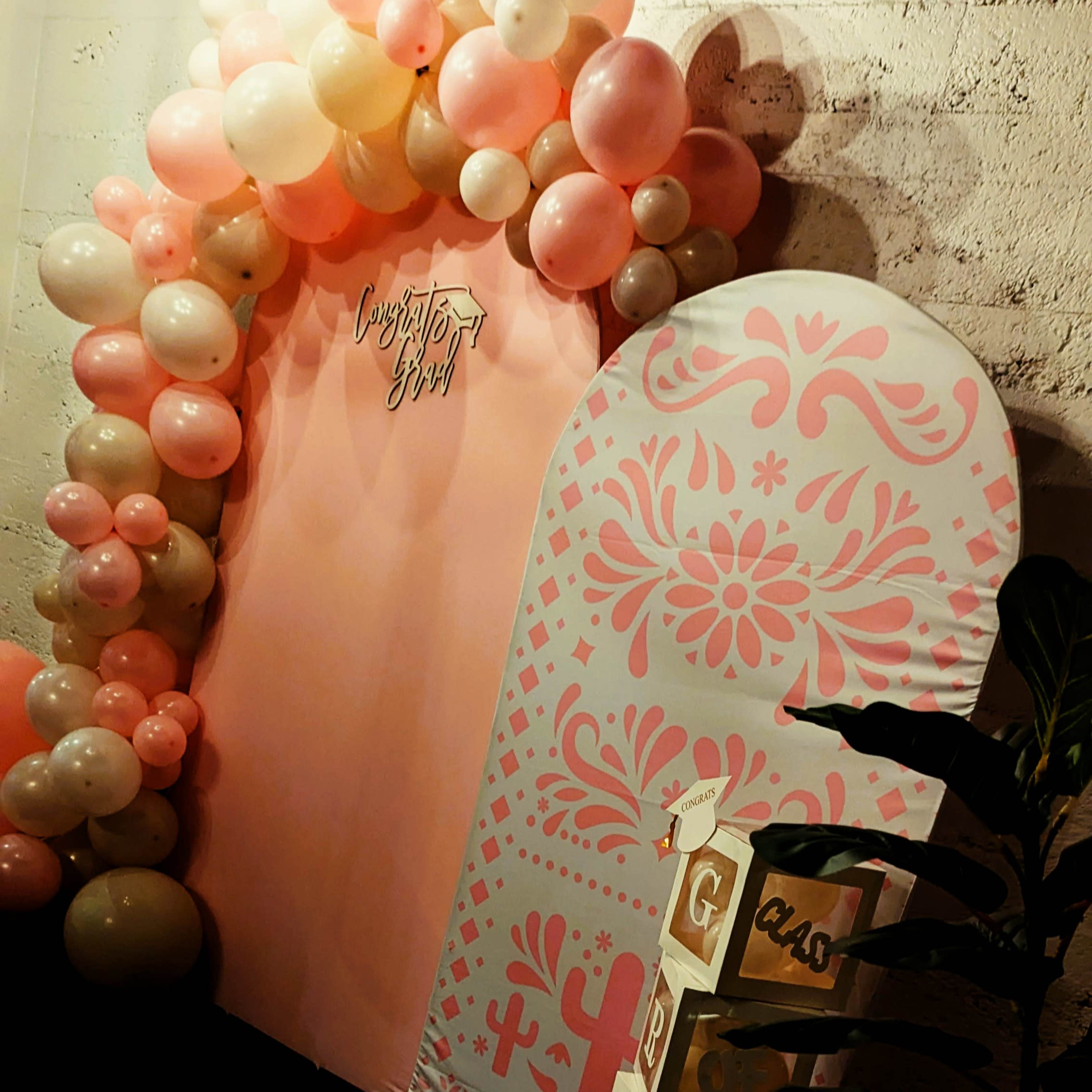 The image shows a decorative setup with a pink balloon arch and a large, patterned display featuring congratulatory text.