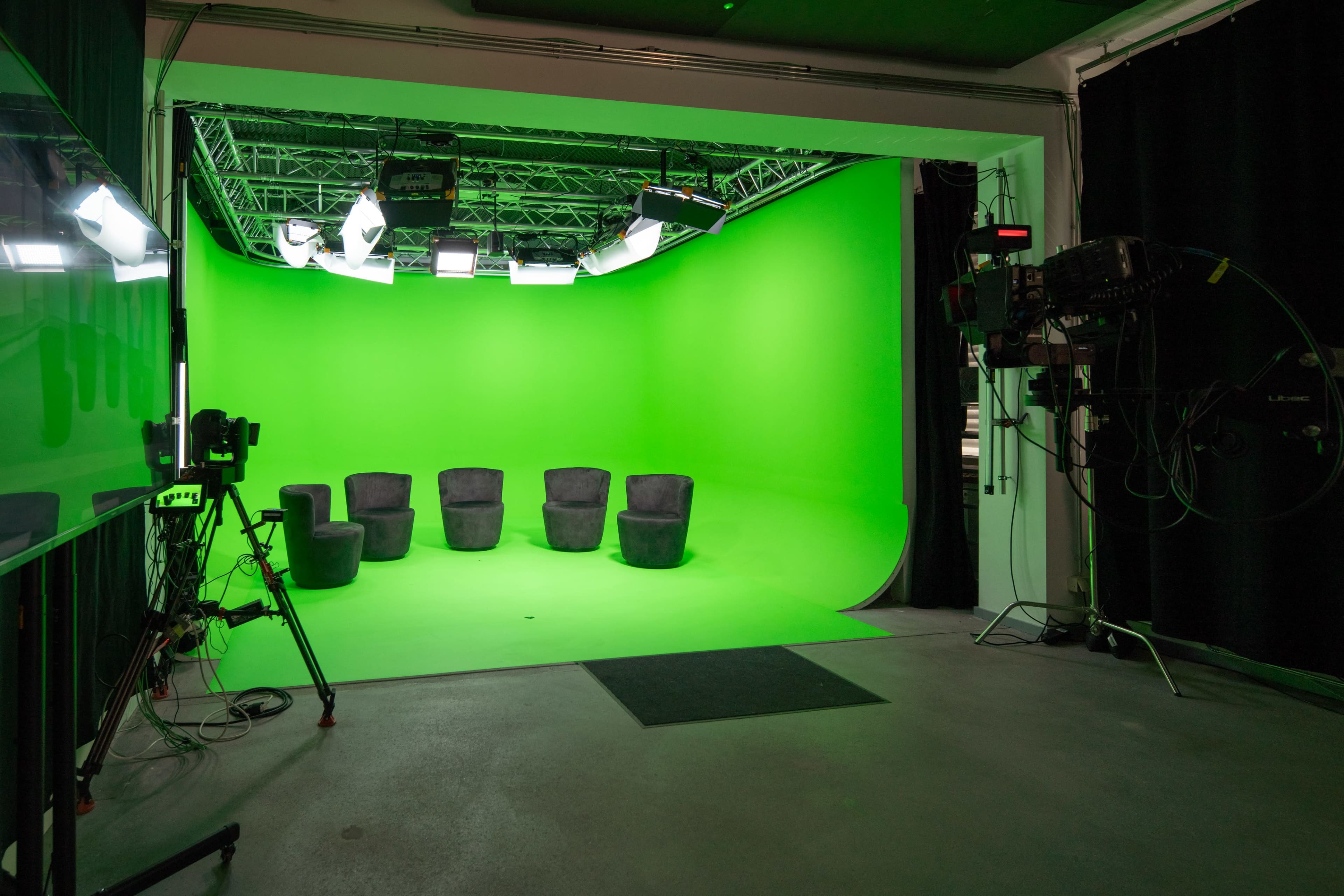 A studio features five black chairs arranged in front of a vibrant green backdrop with lights and cameras set up nearby.