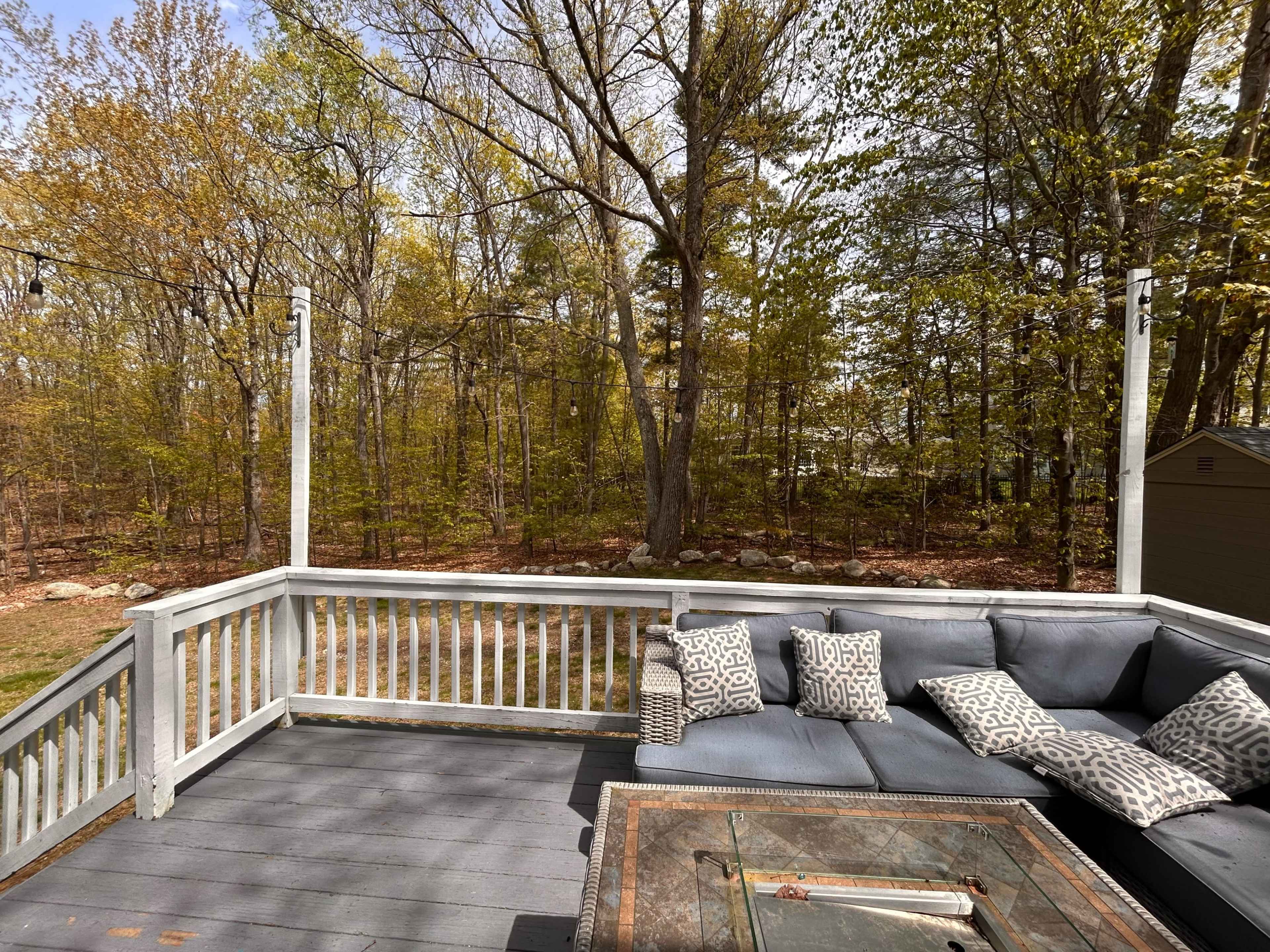 A deck featuring outdoor seating with cushions overlooks a wooded area with trees starting to turn yellow and orange.