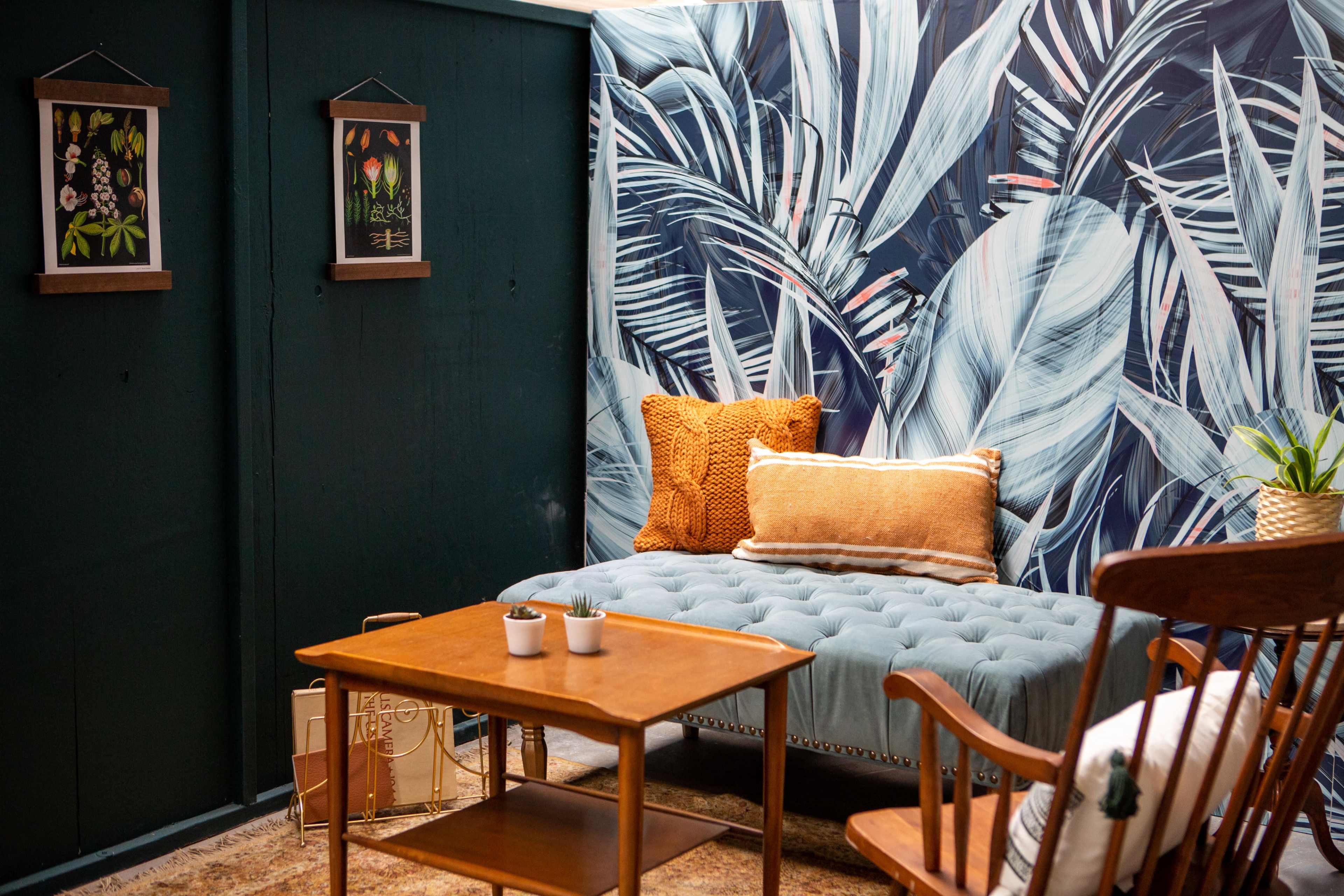 A cozy sitting area featuring a blue tufted couch, a wooden table with plants, and botanical-themed wall art against a backdrop of dark green and leaf-patterned wallpaper.