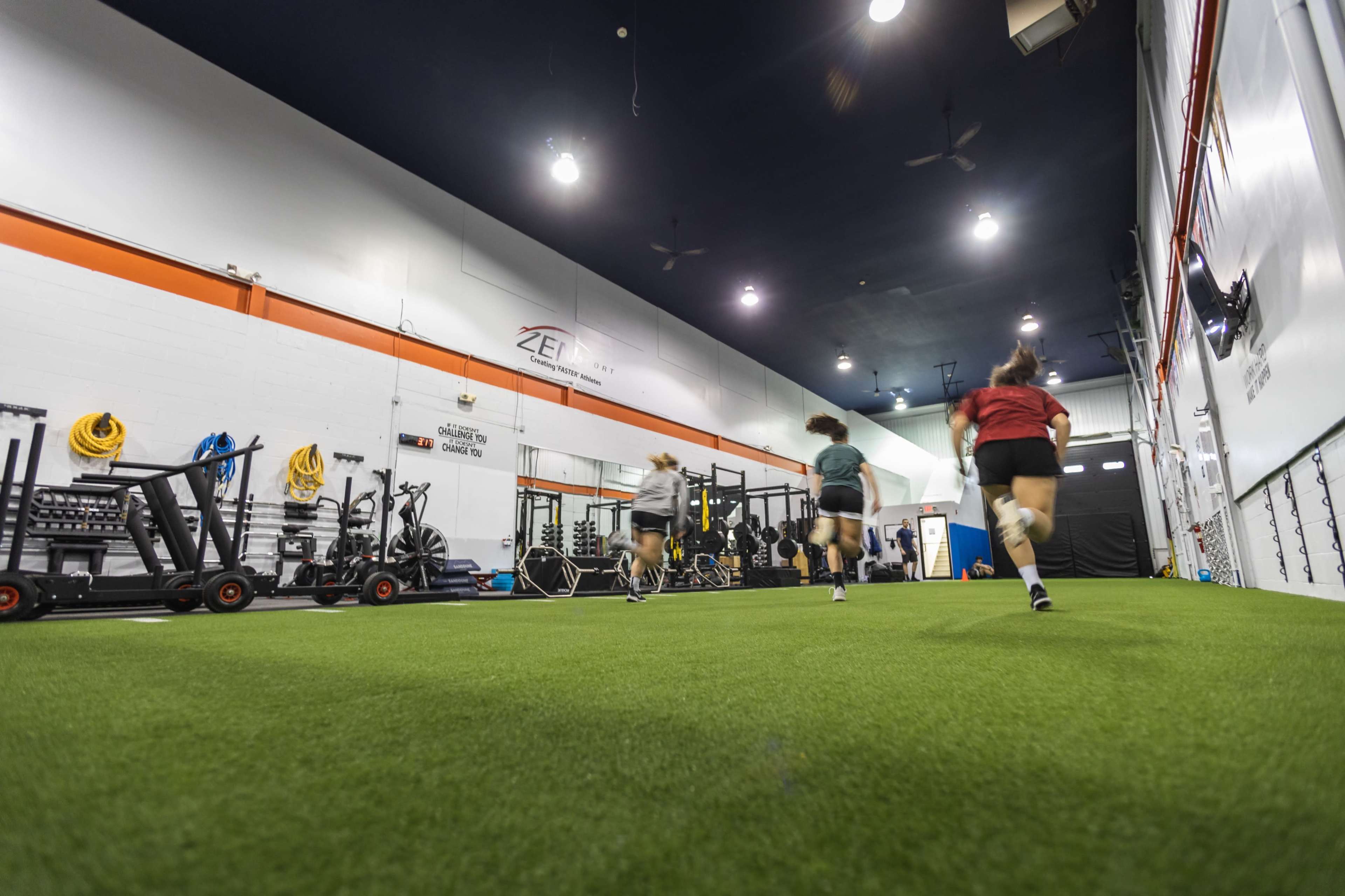 A group of four athletes runs on a turf surface inside a gym equipped with fitness equipment along the walls.