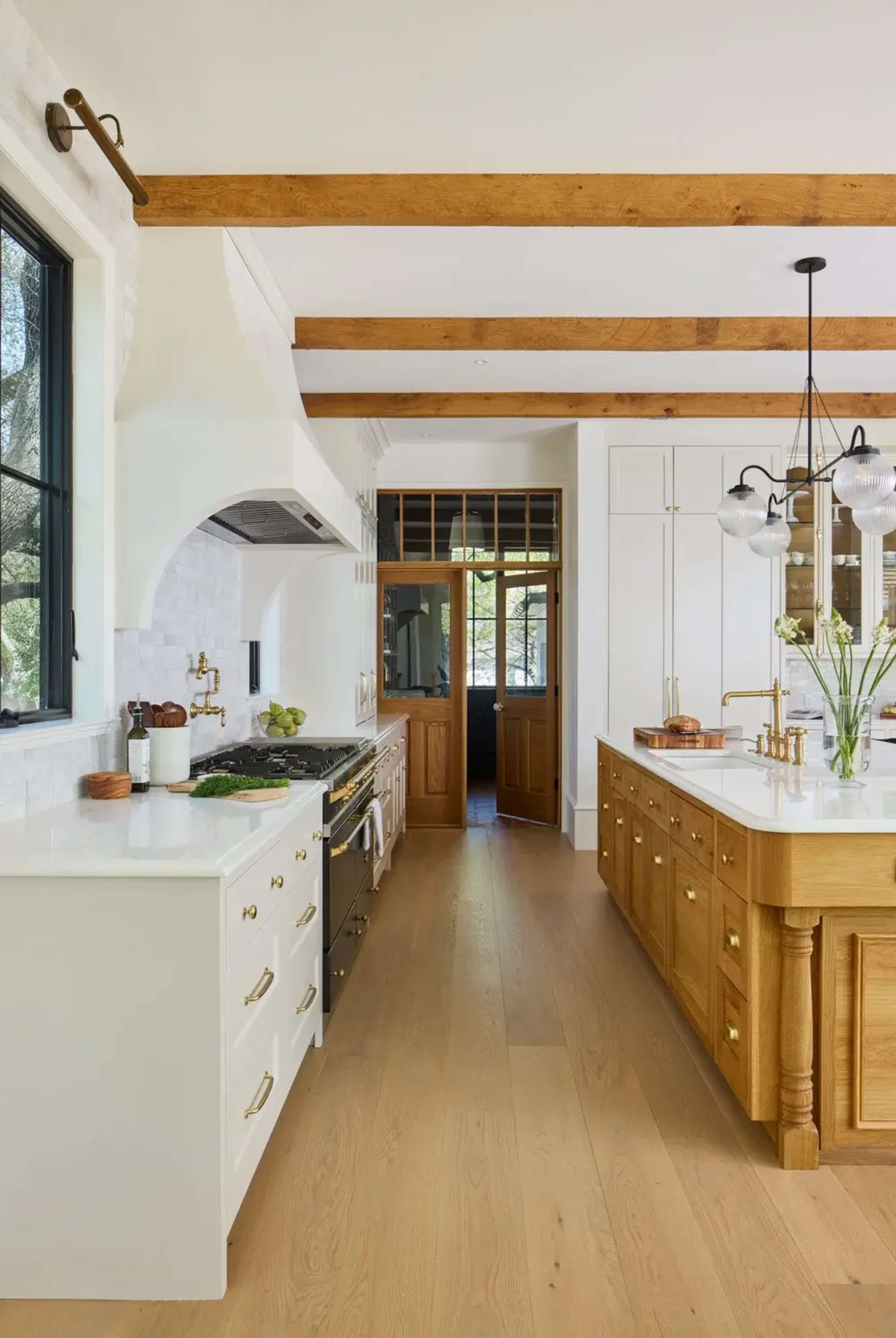 The image depicts a modern kitchen featuring white cabinetry, a large wooden island with brass hardware, and exposed wooden beams overhead.