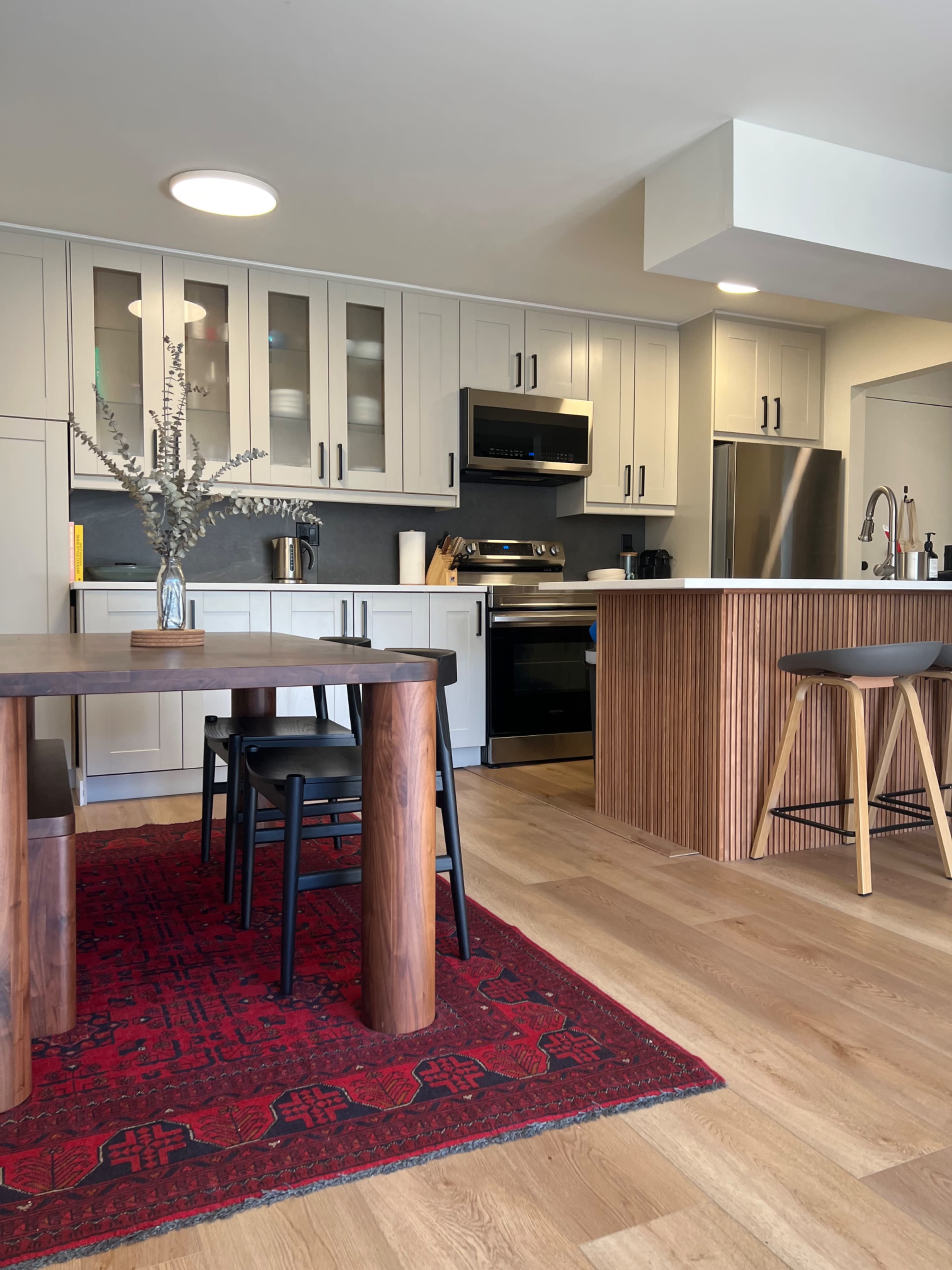 A modern kitchen features stainless steel appliances, white cabinetry, and a wooden dining table on a red patterned rug.