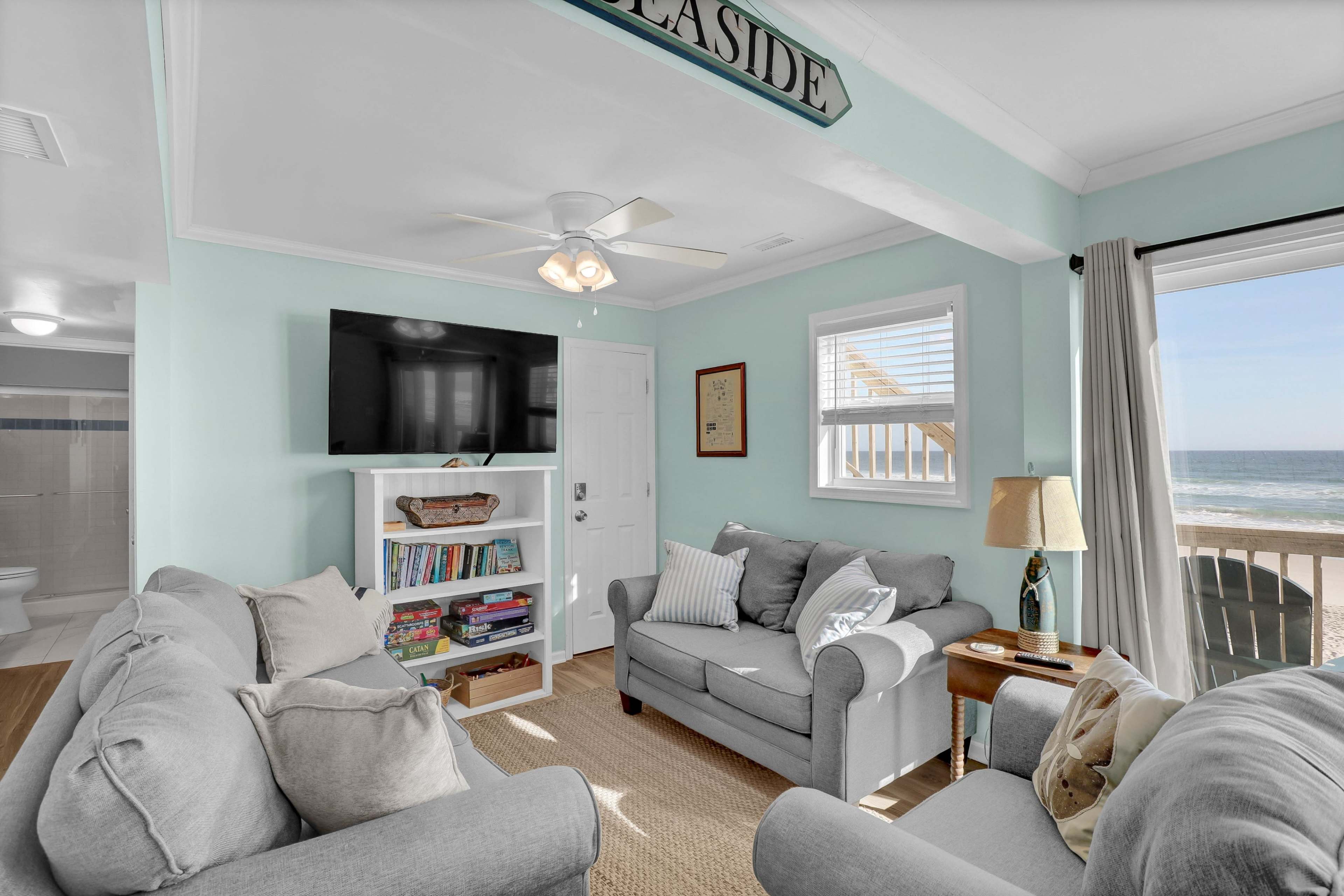 The living area features a light blue wall, a ceiling fan, a large television, and a view of the ocean through a window.