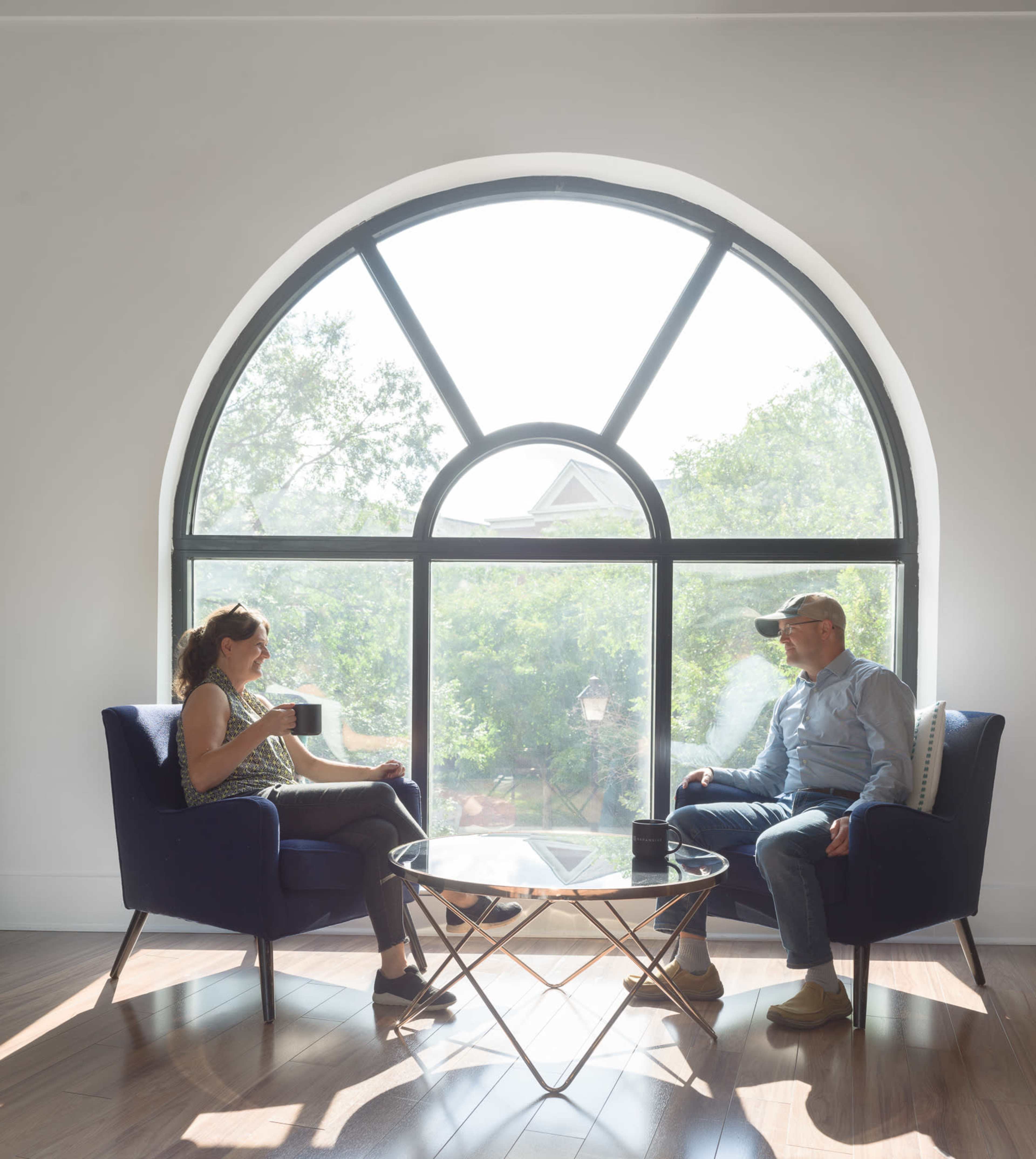 Two people sit in individual chairs by a large arched window, engaged in conversation, with a table between them and greenery visible outside.