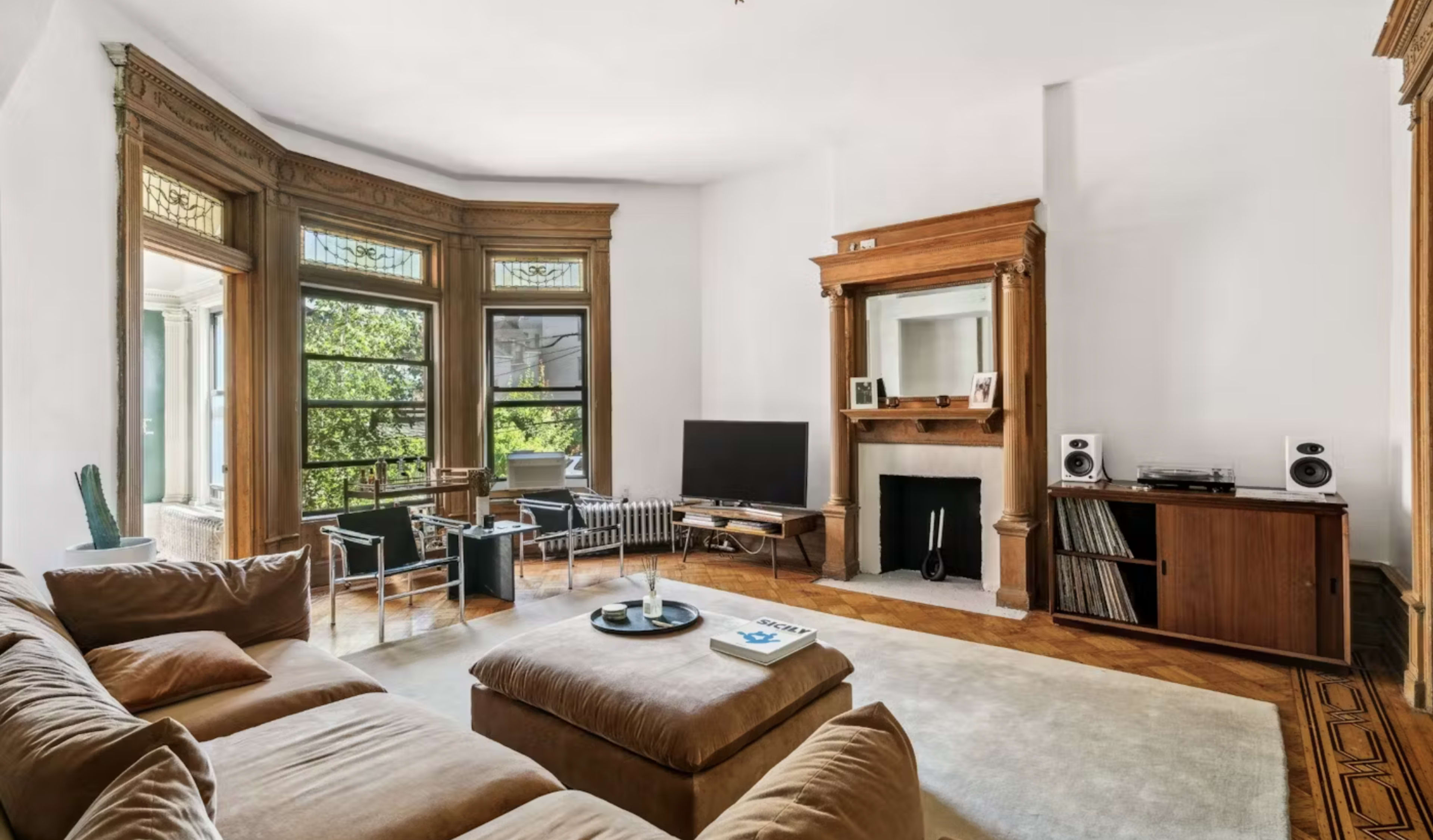 The image shows a living room with a large sectional sofa, a coffee table, a television mounted on the wall, and a fireplace flanked by audio equipment.