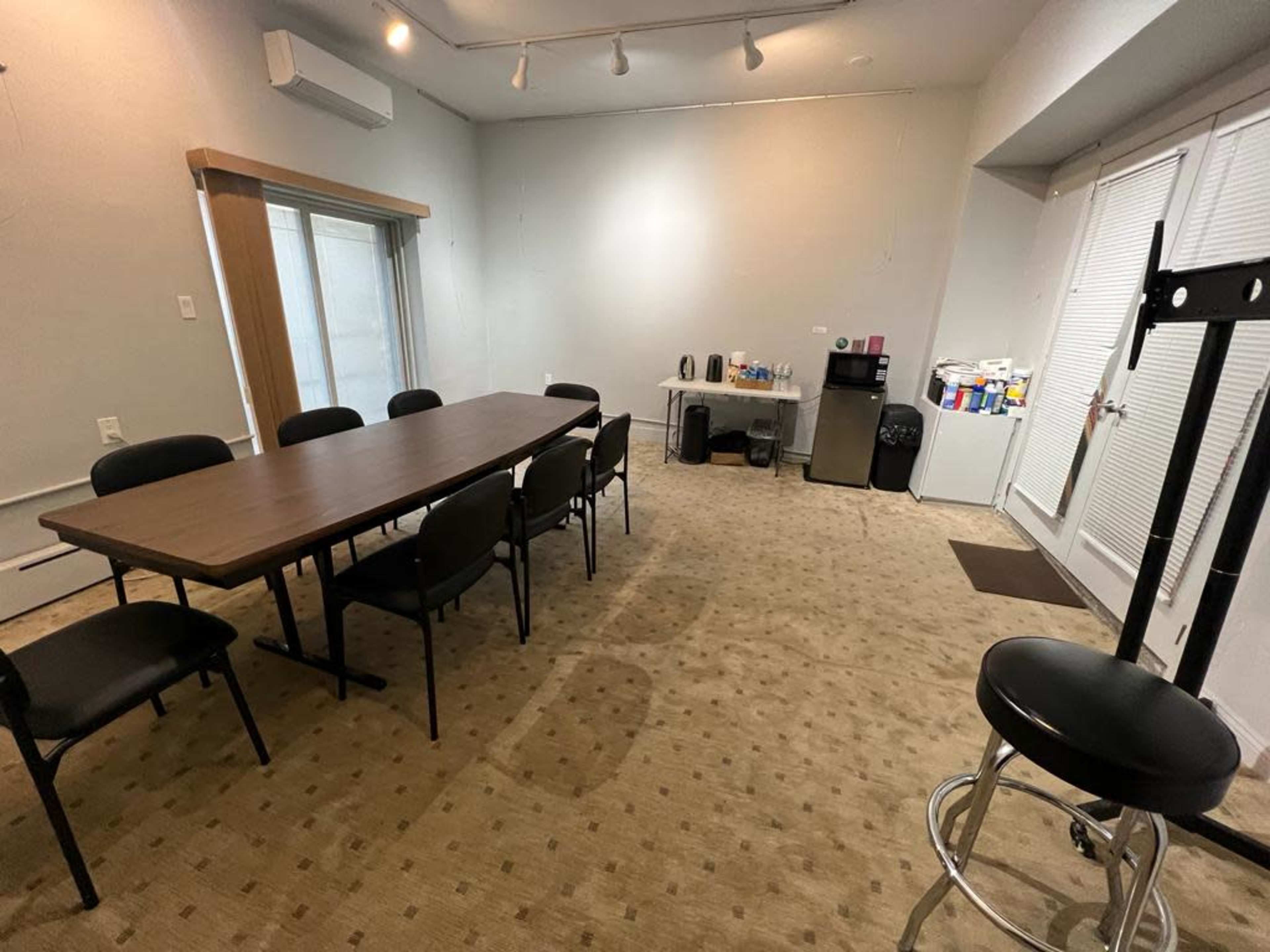 A meeting room with a long table surrounded by chairs, a kitchenette area in the background, and a small bar stool in the corner.