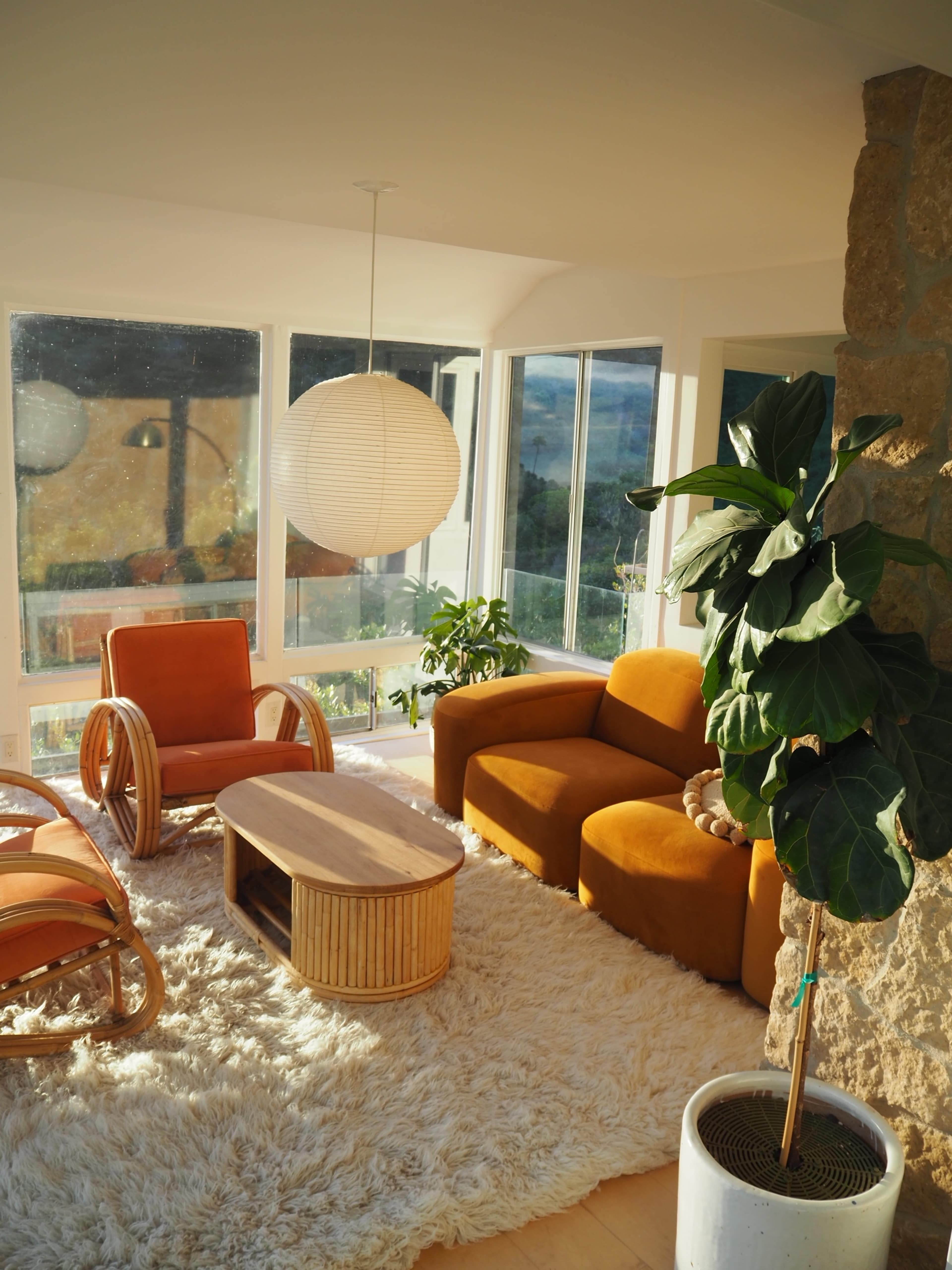 The image shows a sunlit living area featuring orange armchairs, a round paper lantern light, a wooden coffee table, and a potted plant next to a stone wall.