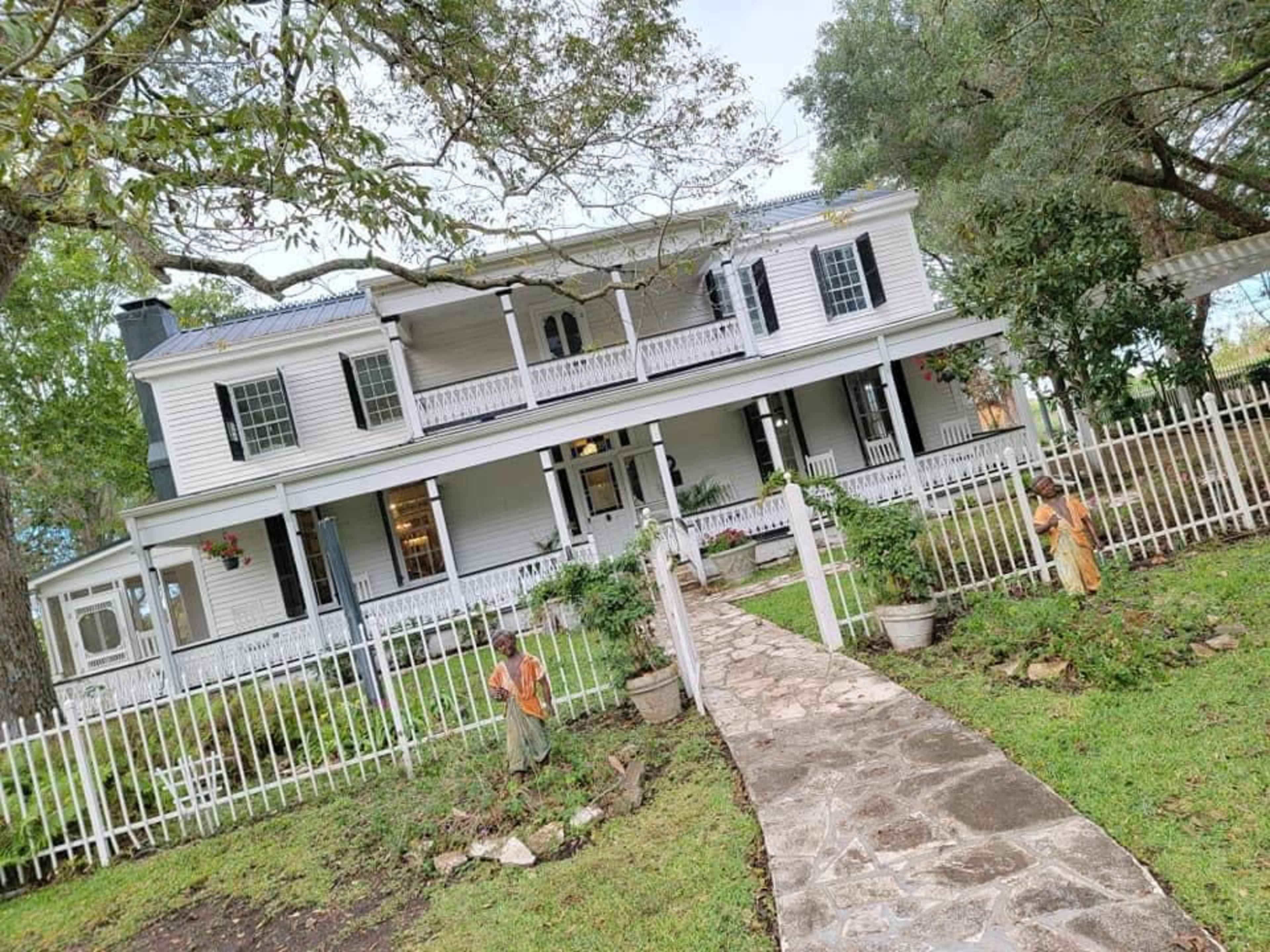 A two-story white house with a front porch and a stone pathway is surrounded by a white picket fence and small garden statues.