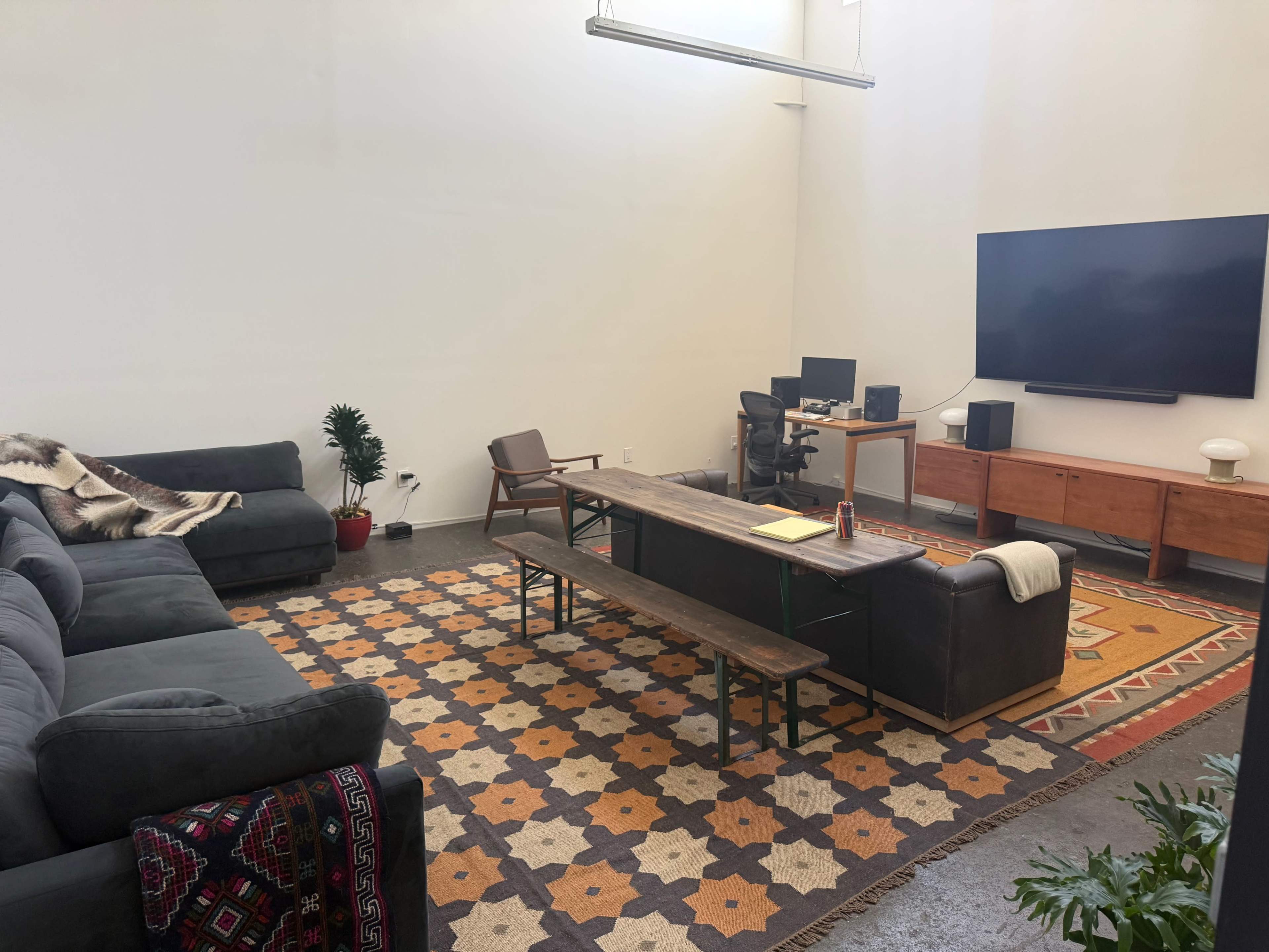 The image shows a spacious living room featuring a large gray sectional sofa, a long wooden coffee table, a wall-mounted TV, and a patterned rug on the floor.