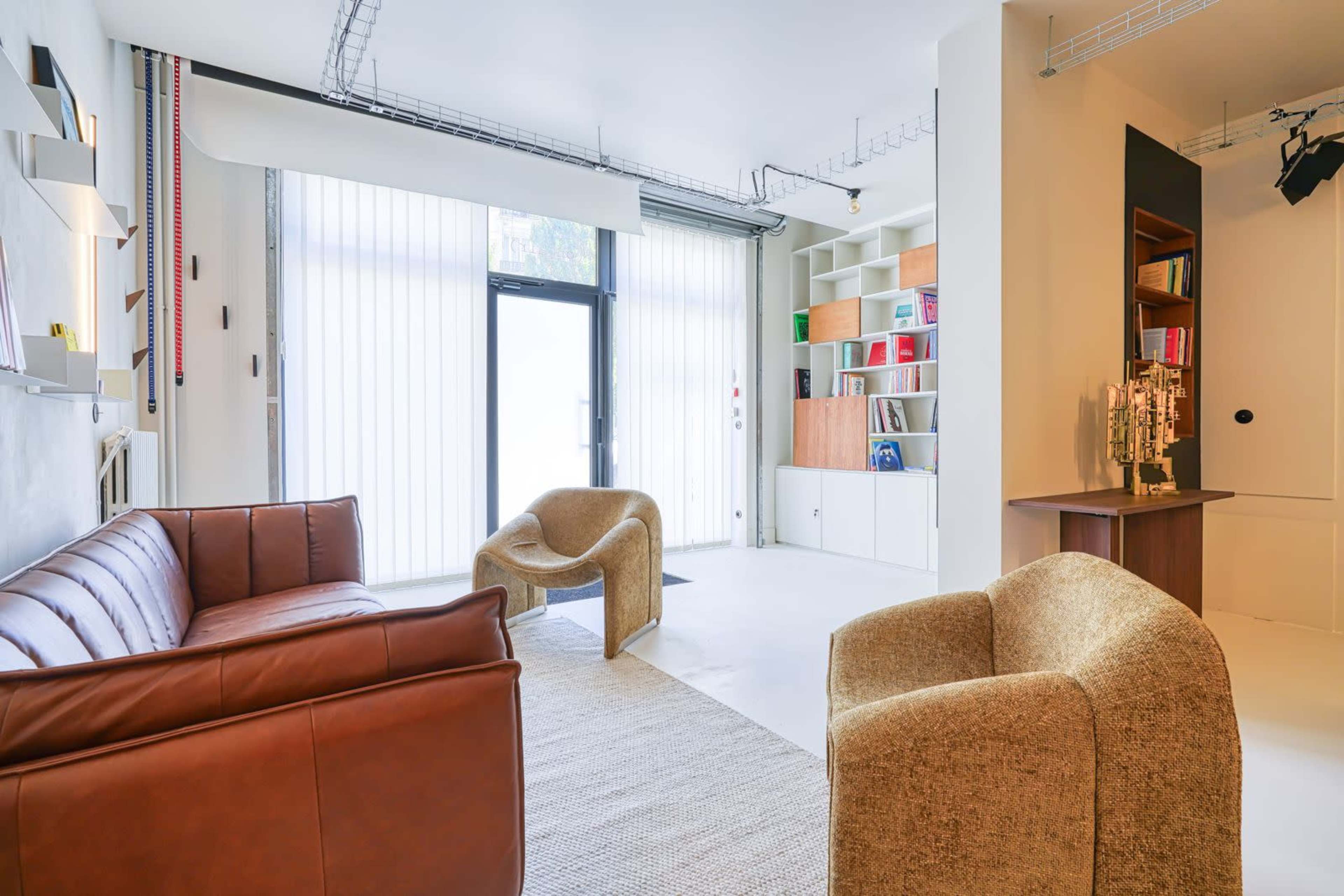 A modern living room features a leather sofa, two fabric chairs, a rug, and shelves filled with books and decorative items.