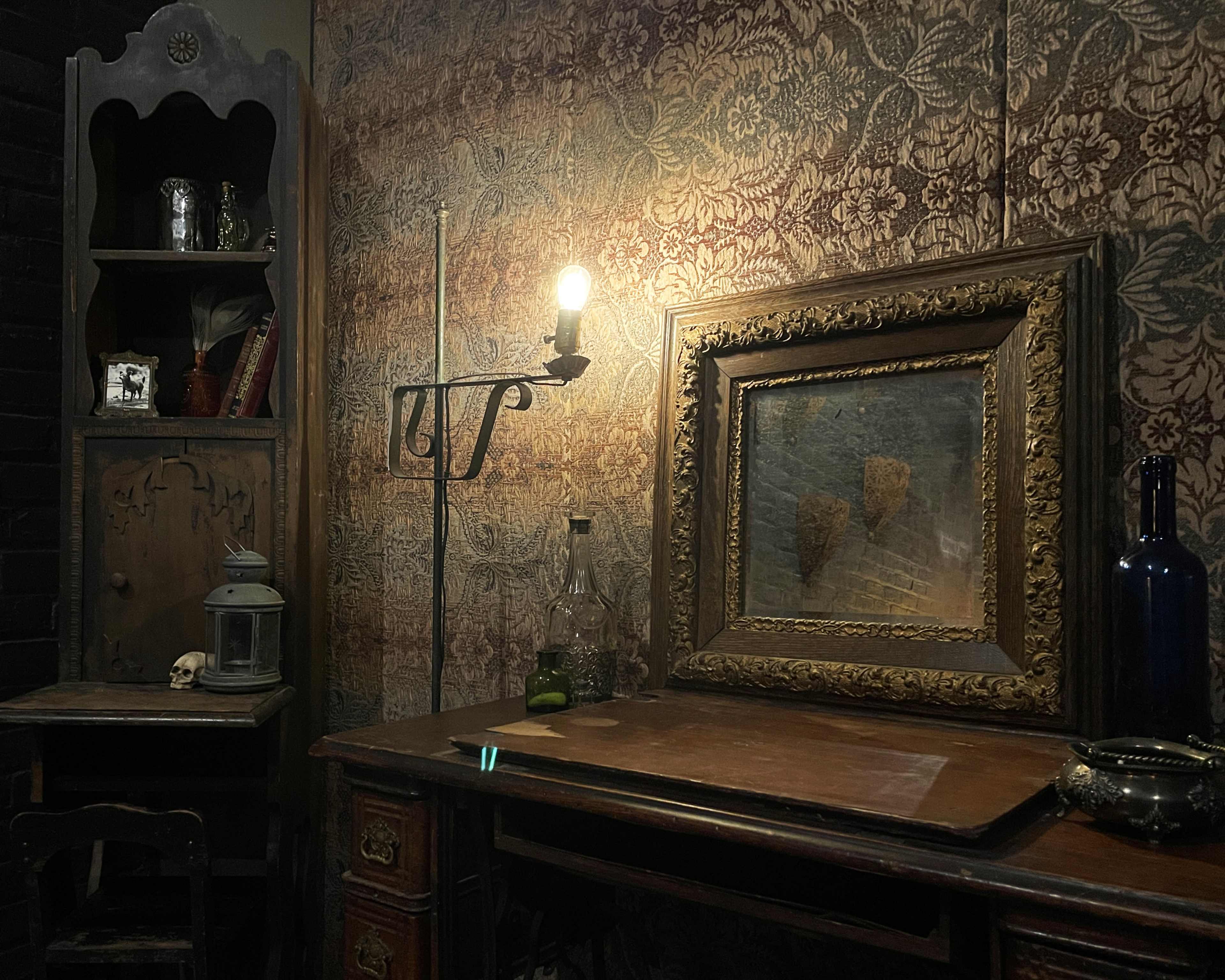 The image shows a dimly lit room with antique furniture, including a wooden desk, a bookcase, and a framed painting against a patterned wallpaper backdrop.