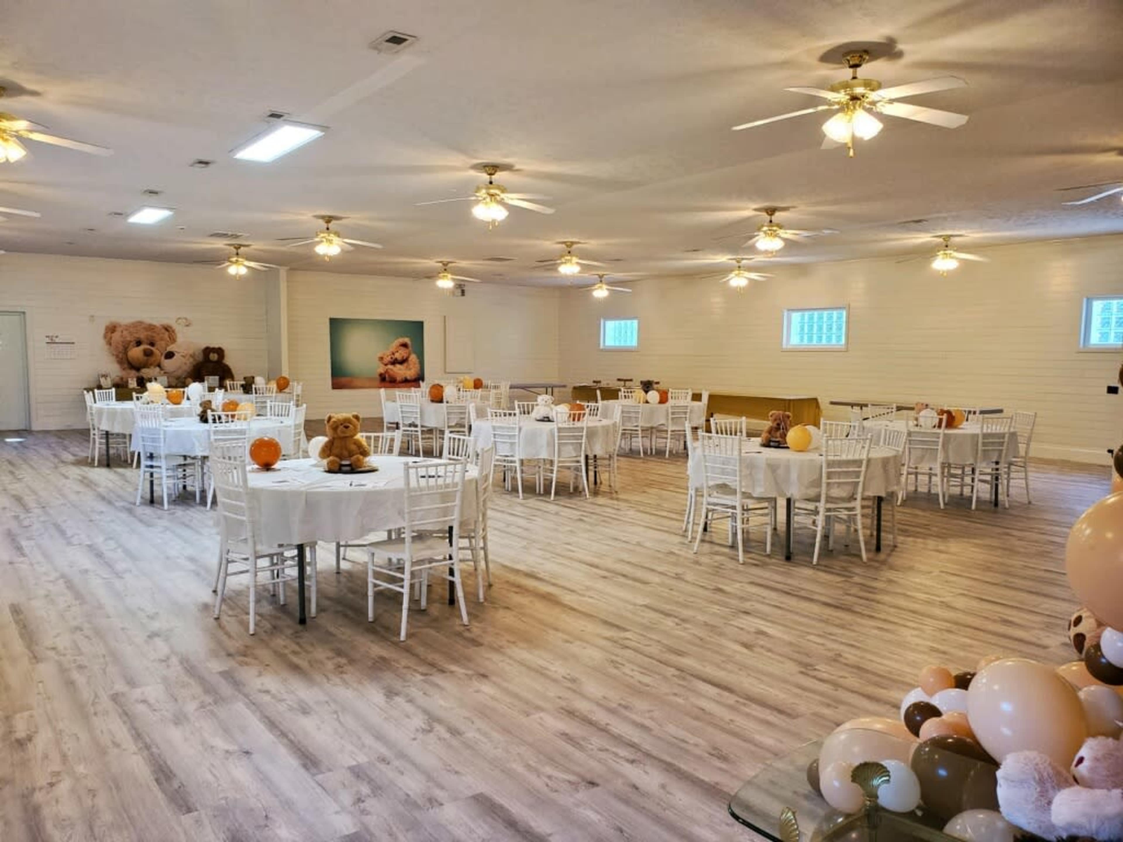 A spacious event room with white tables set for an occasion, surrounded by decorative teddy bears and balloons.