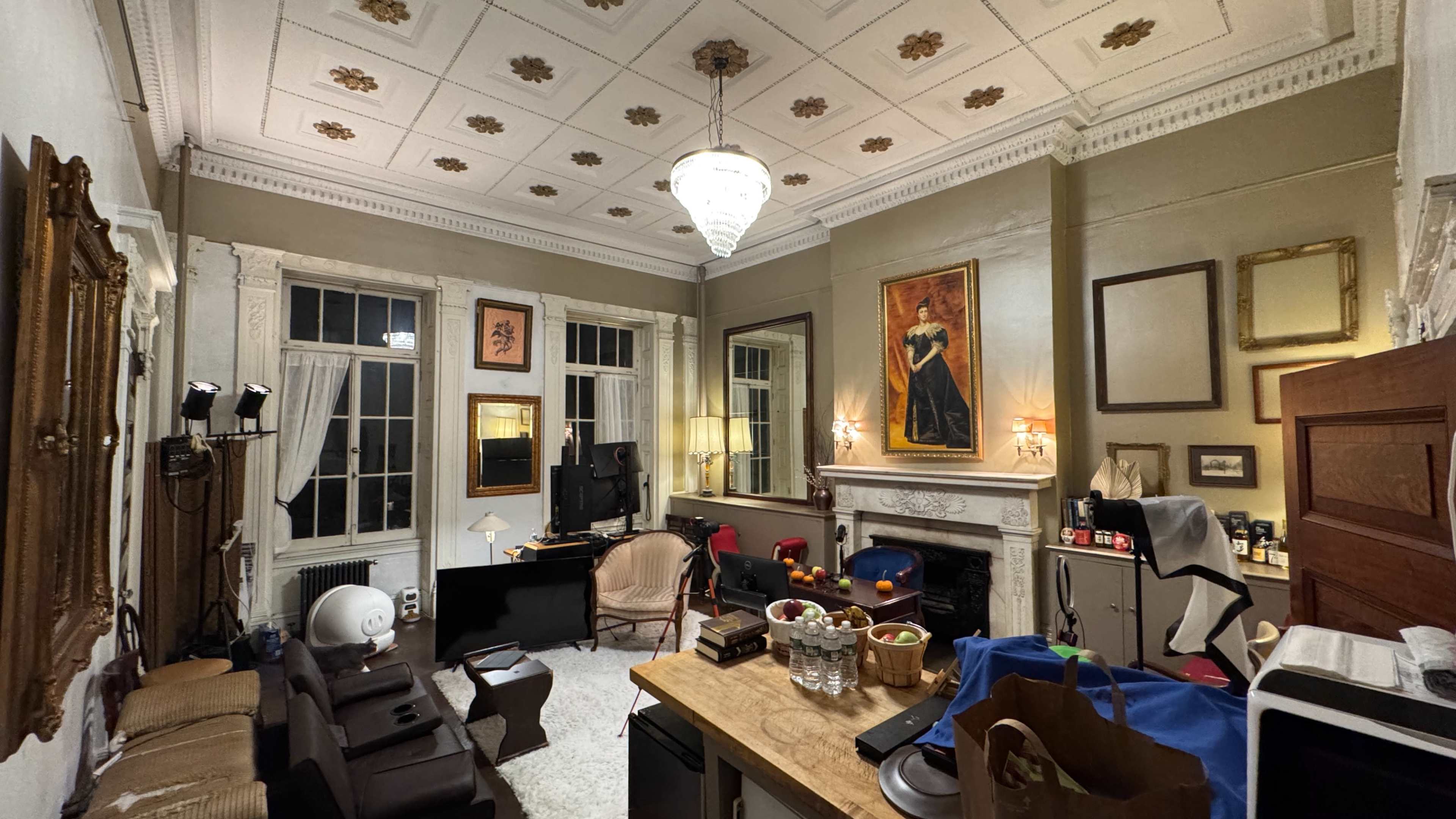 The image shows a spacious room with high ceilings, featuring a mix of furniture, a computer workstation, and various items scattered around, including a table with several containers.