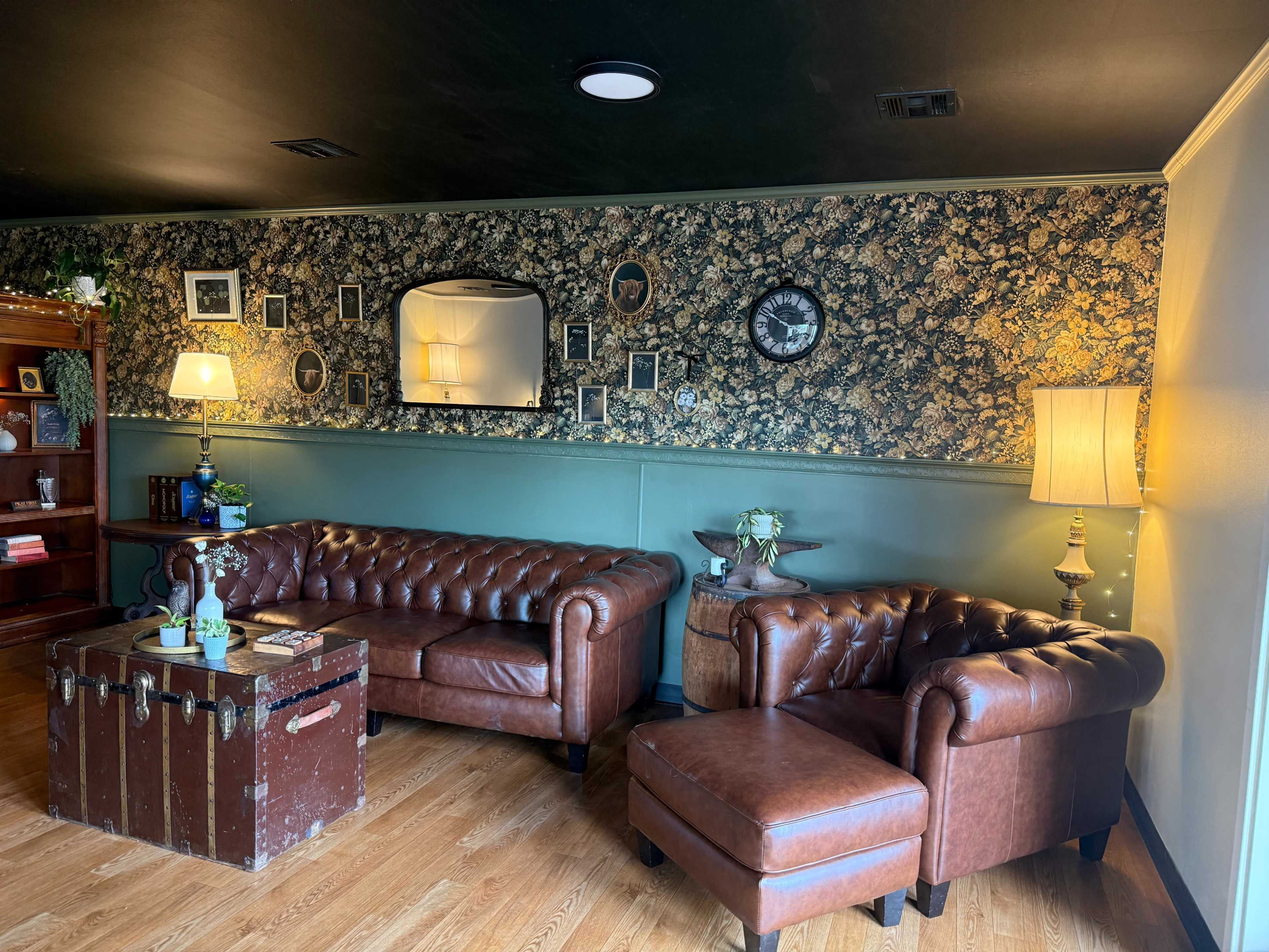 The image shows a cozy living area with a brown leather chesterfield sofa, an armchair, a side table, and vintage decor on the walls, including framed pictures and lamps.