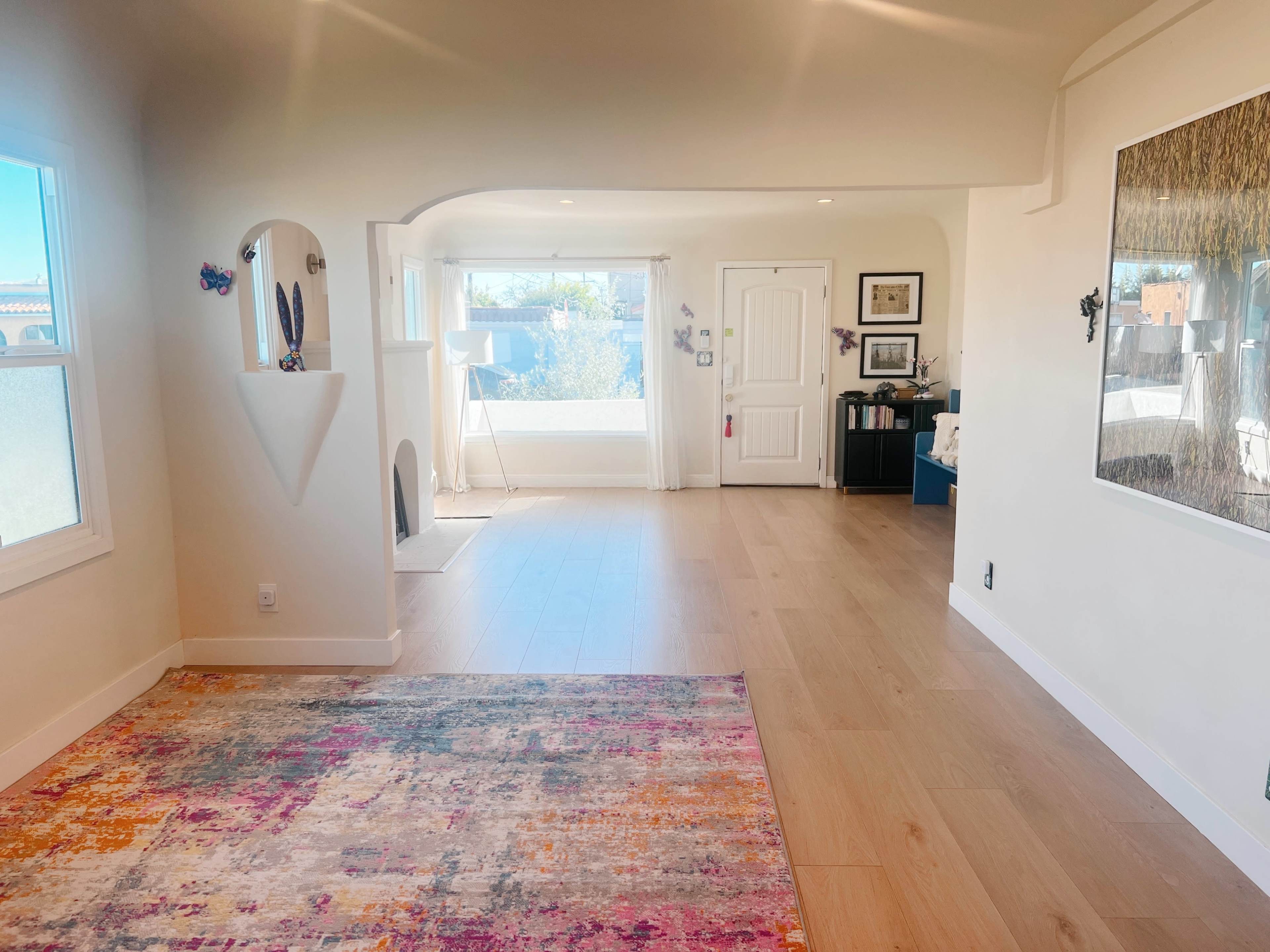 The image shows a bright, open interior space featuring a living area with large windows, light wooden flooring, and a colorful area rug.