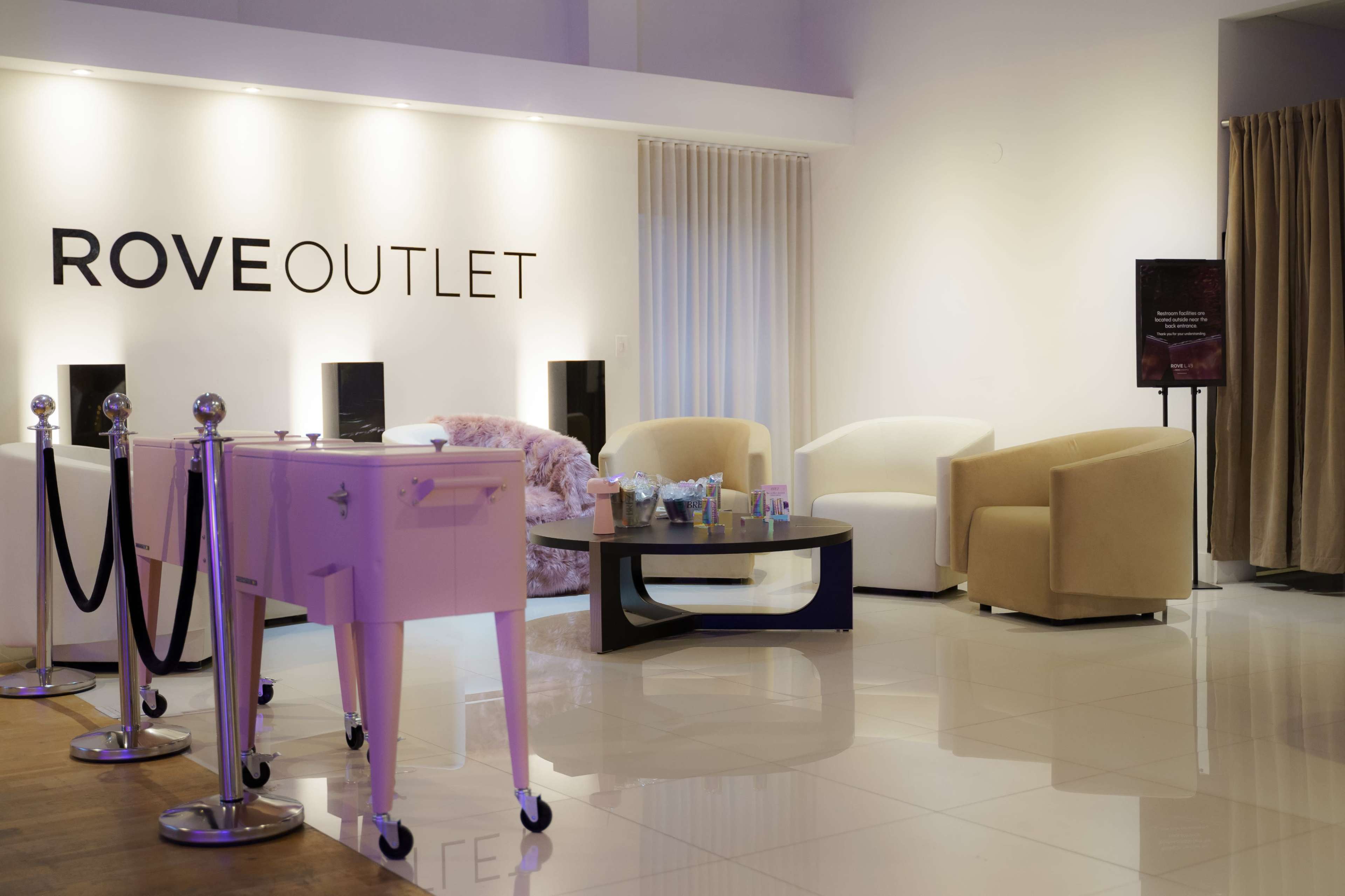 The image shows a modern lounge area with comfortable seating, a coffee table, and a reception setup at the ROVE OUTLET.