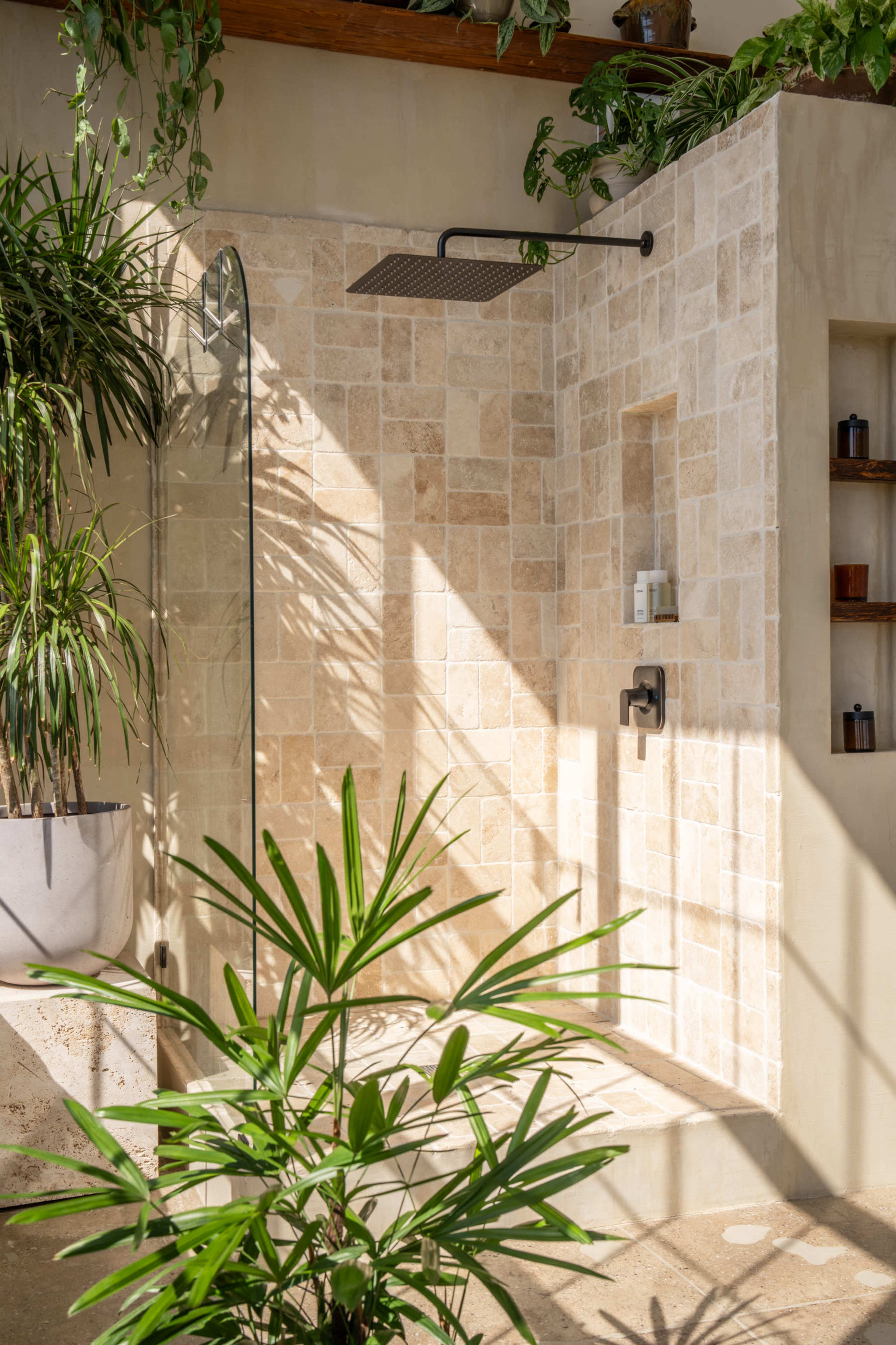 The image shows a modern shower area featuring natural stone walls, a large overhead shower head, and potted plants.