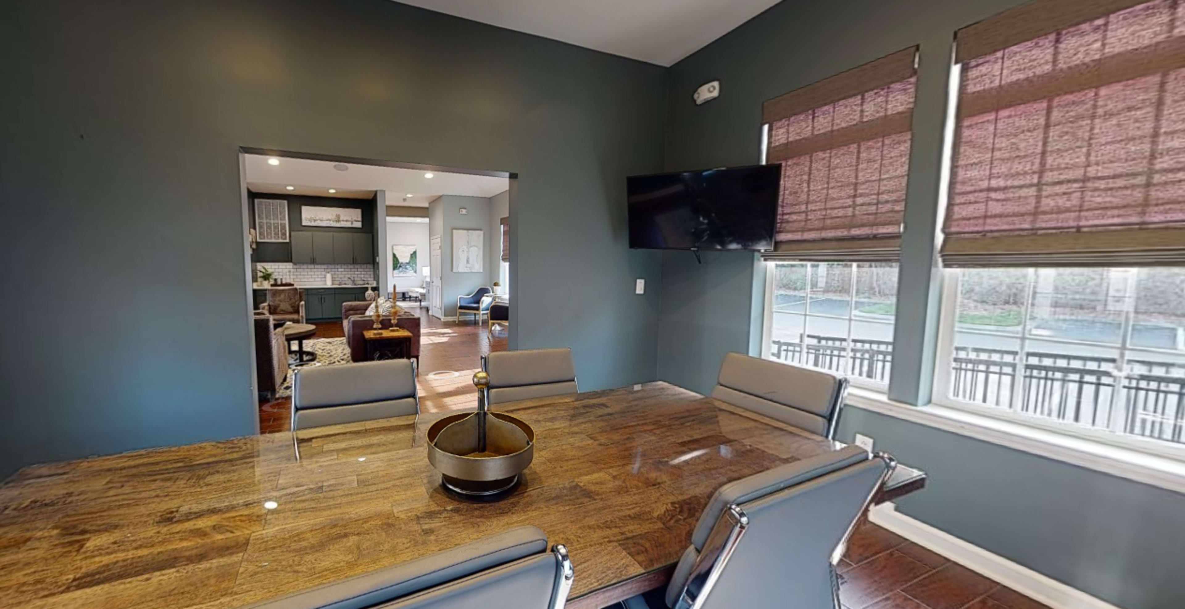 A dining room features a large wooden table with eight chairs, a bowl centerpiece, and a wall-mounted television, with windows overlooking a patio area.