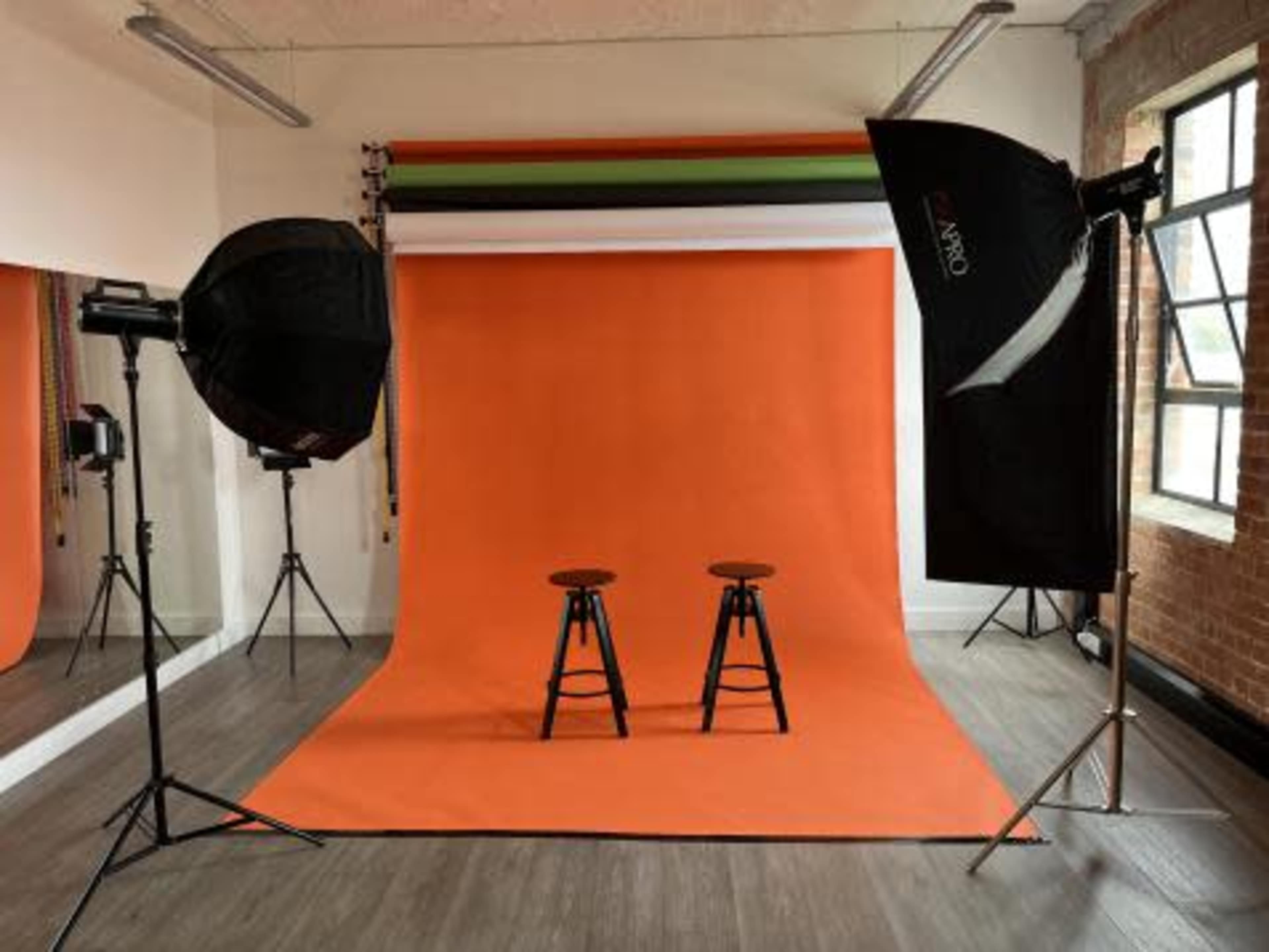 A photography studio with two wooden stools positioned in front of an orange backdrop, surrounded by lighting equipment.