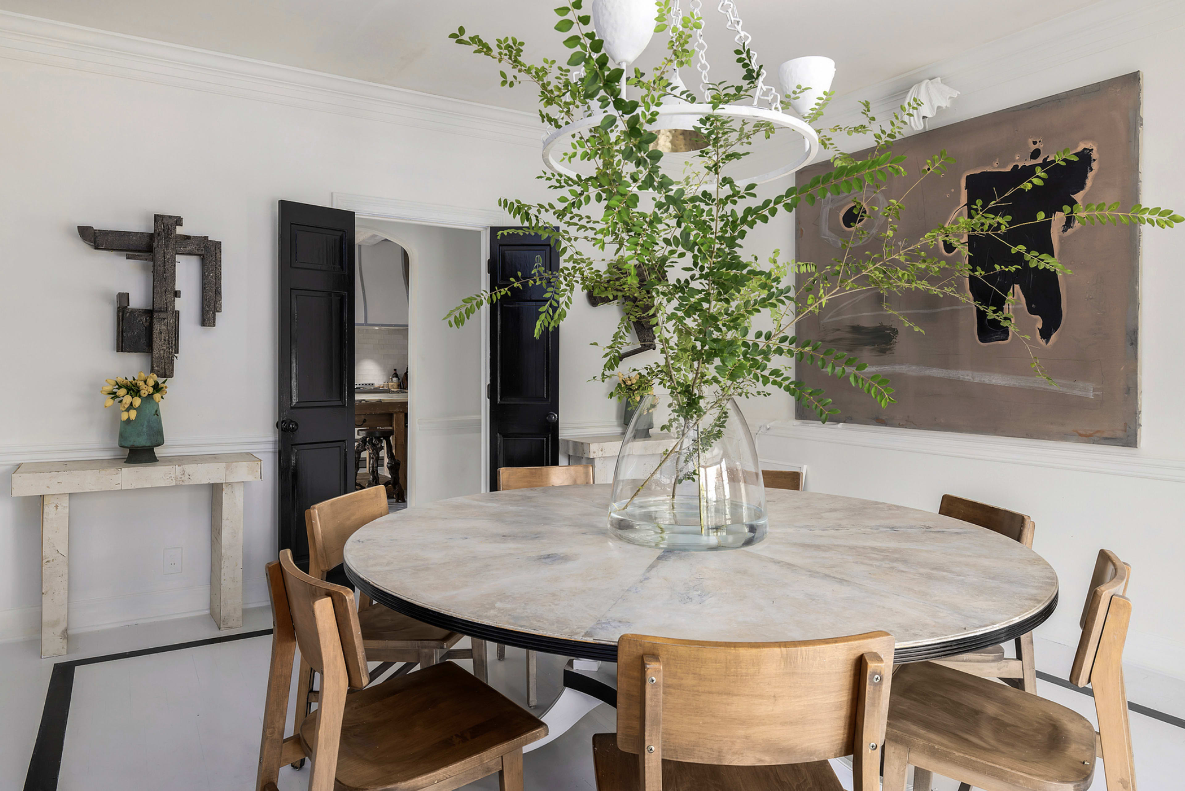 A round wooden dining table is set with a large glass vase filled with greenery, surrounded by wooden chairs in a bright, minimalist dining area featuring abstract art on the walls.