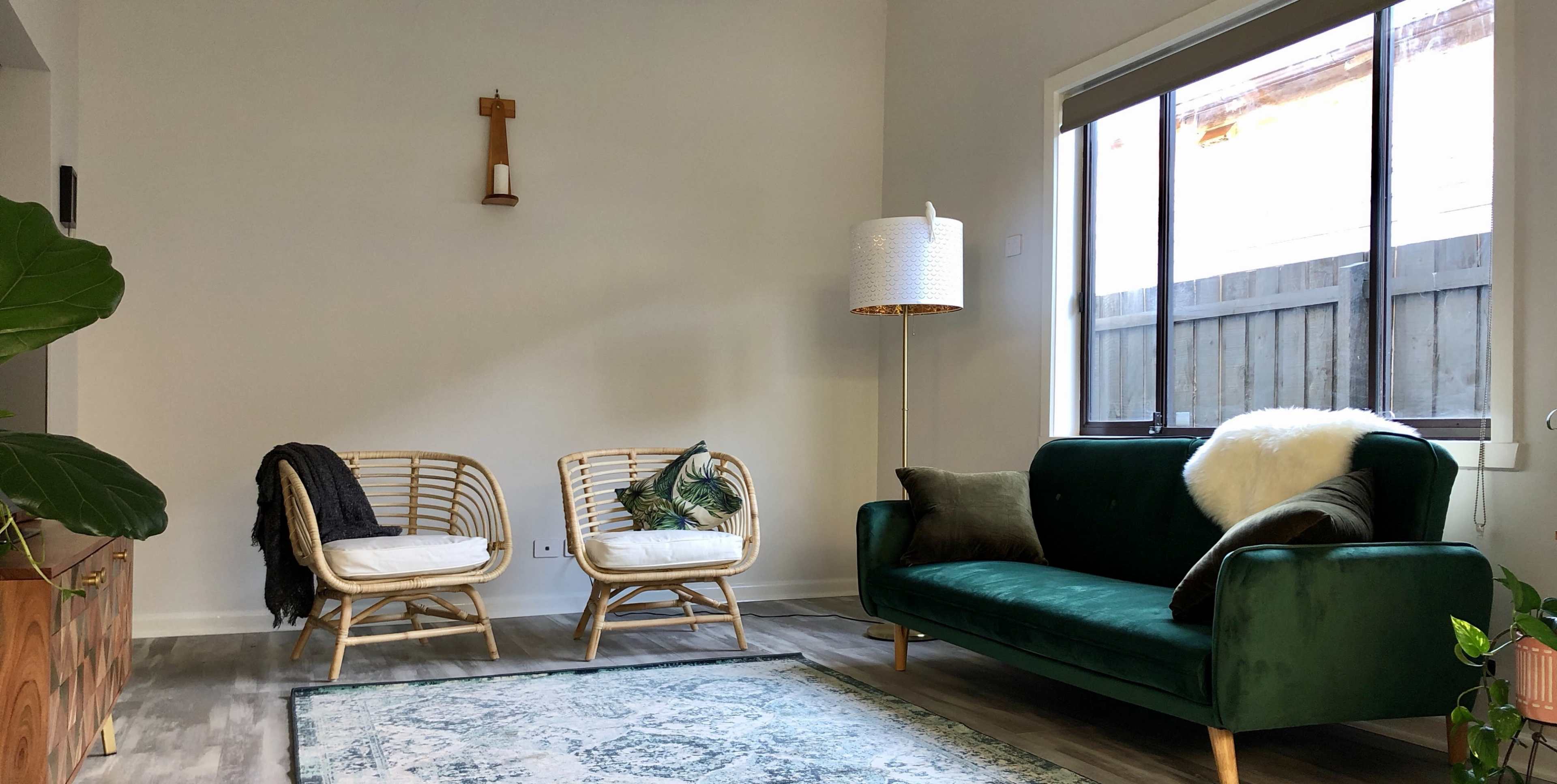 The image shows a living room with two rattan chairs, a green sofa, a floor lamp, and a decorative rug, all arranged against a light-colored wall and a window.