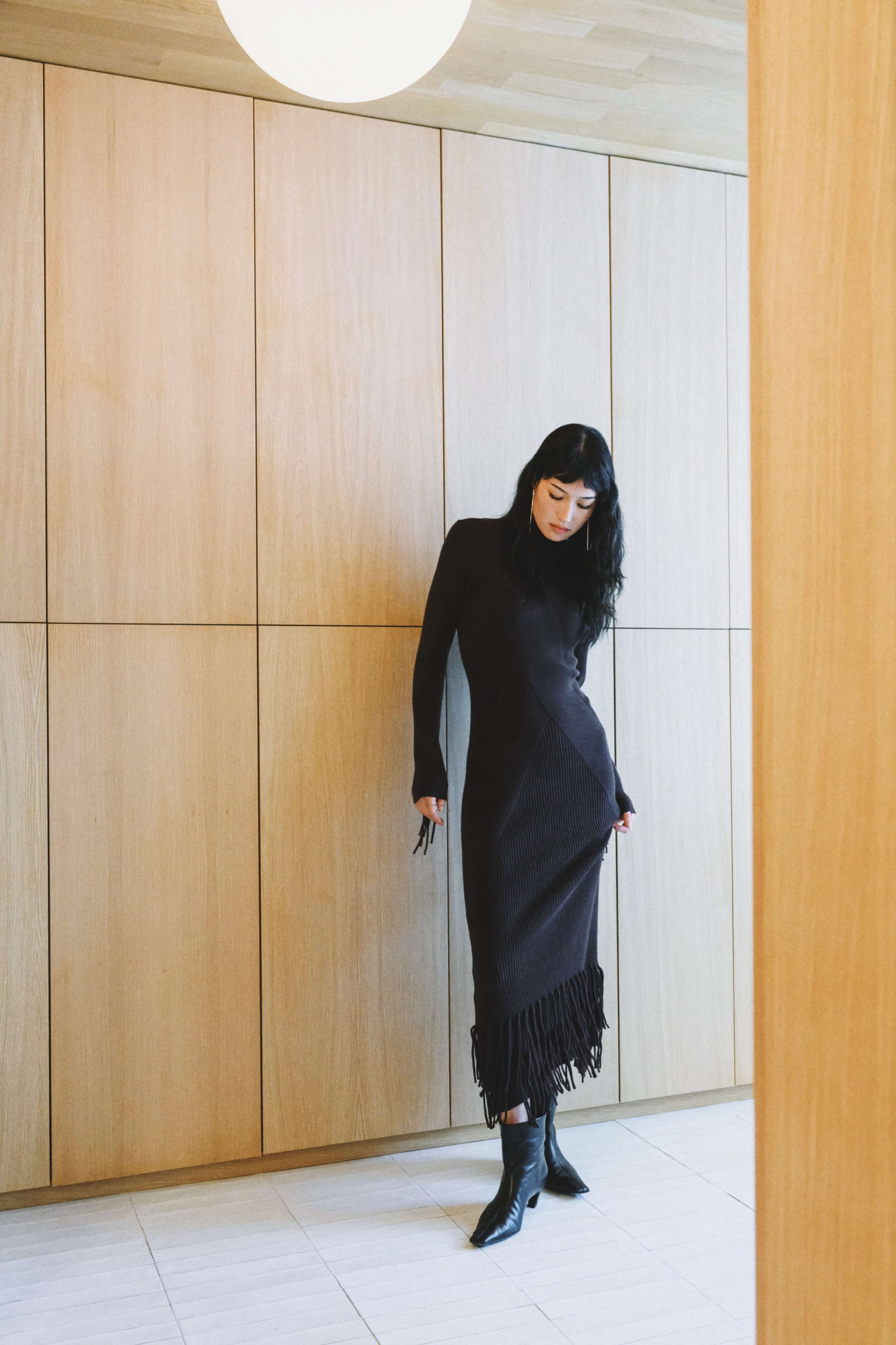 A woman wearing a black dress with fringe stands in a room with wooden paneling and a round light fixture.