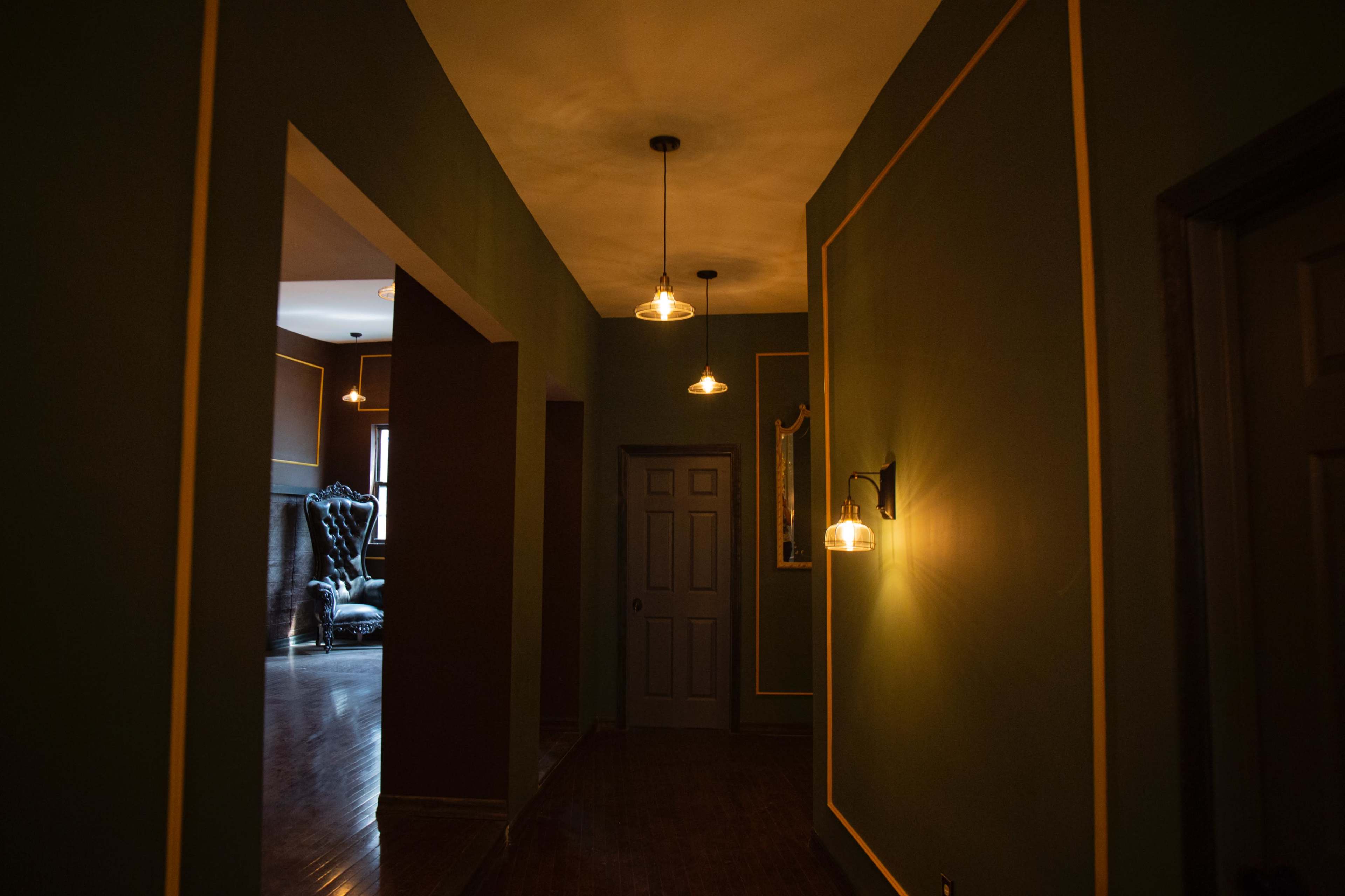 A dimly lit hallway features green walls with gold trim and vintage-style pendant lights, leading to another room with a dark door.