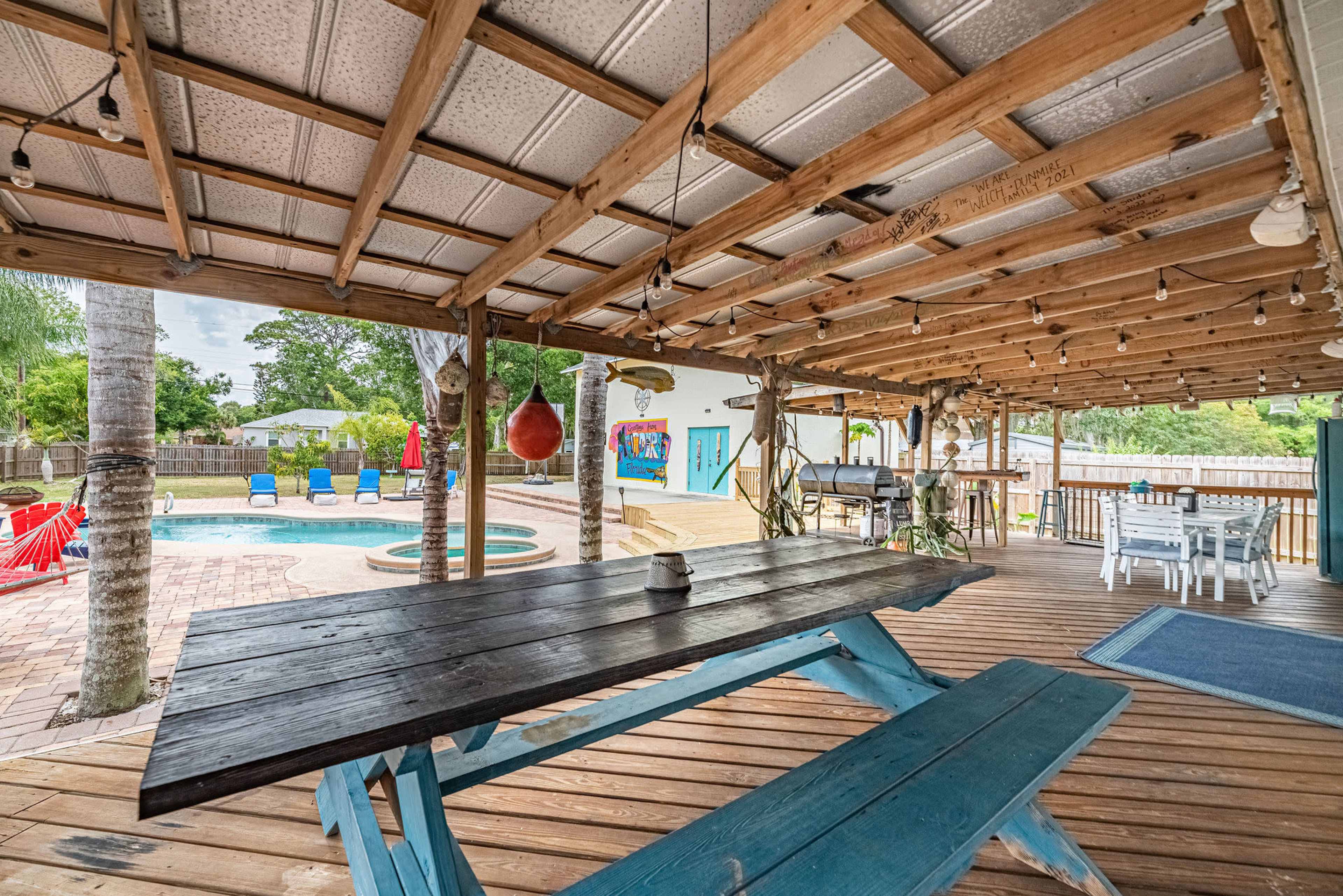 A wooden deck with a large picnic table, overlooking a pool area surrounded by lounge chairs and colorful decorations.