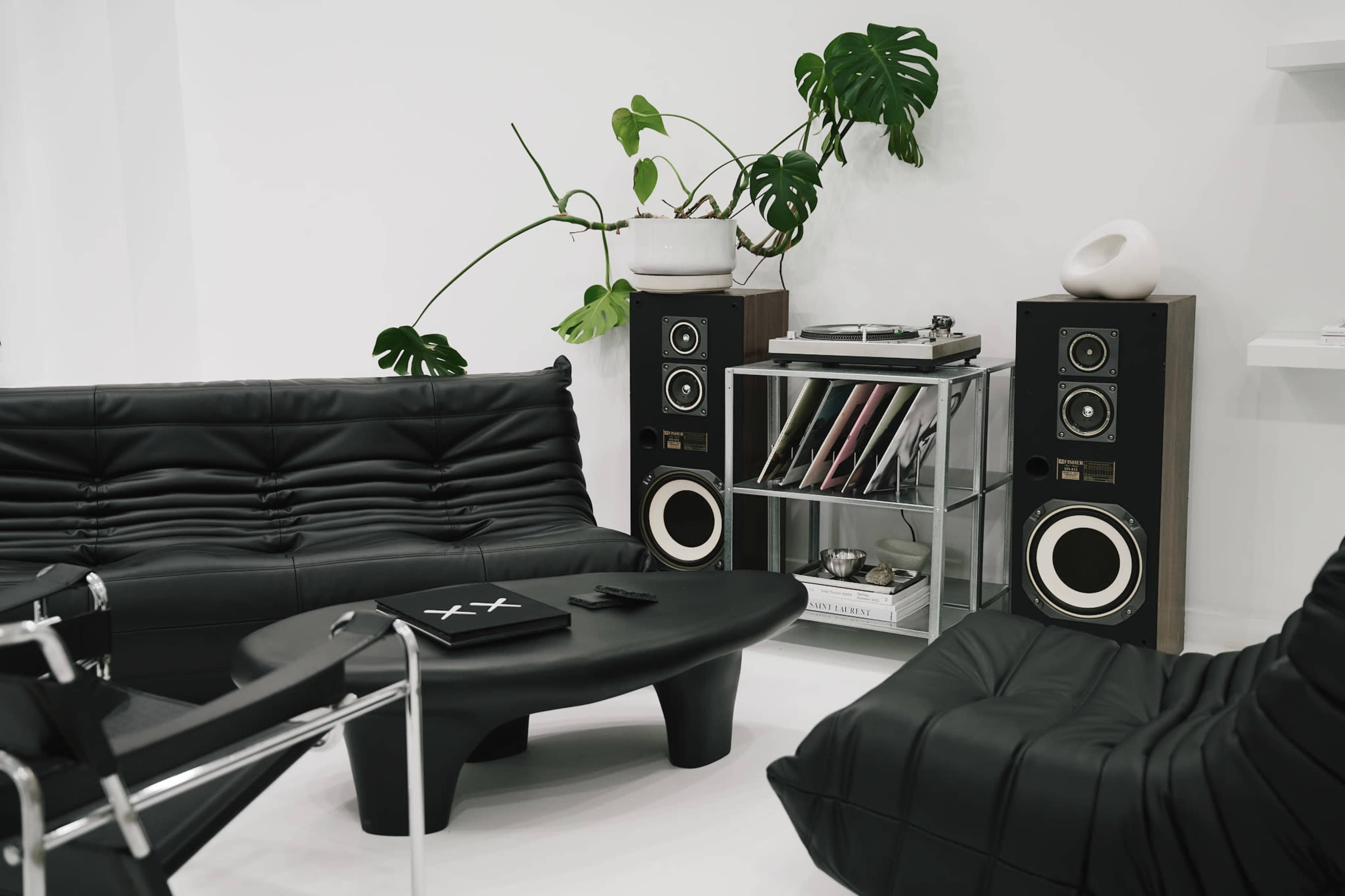 The image shows a minimalist living room with black sofas, a round coffee table, large speakers, a turntable, and a shelf containing vinyl records, all set against a white wall.