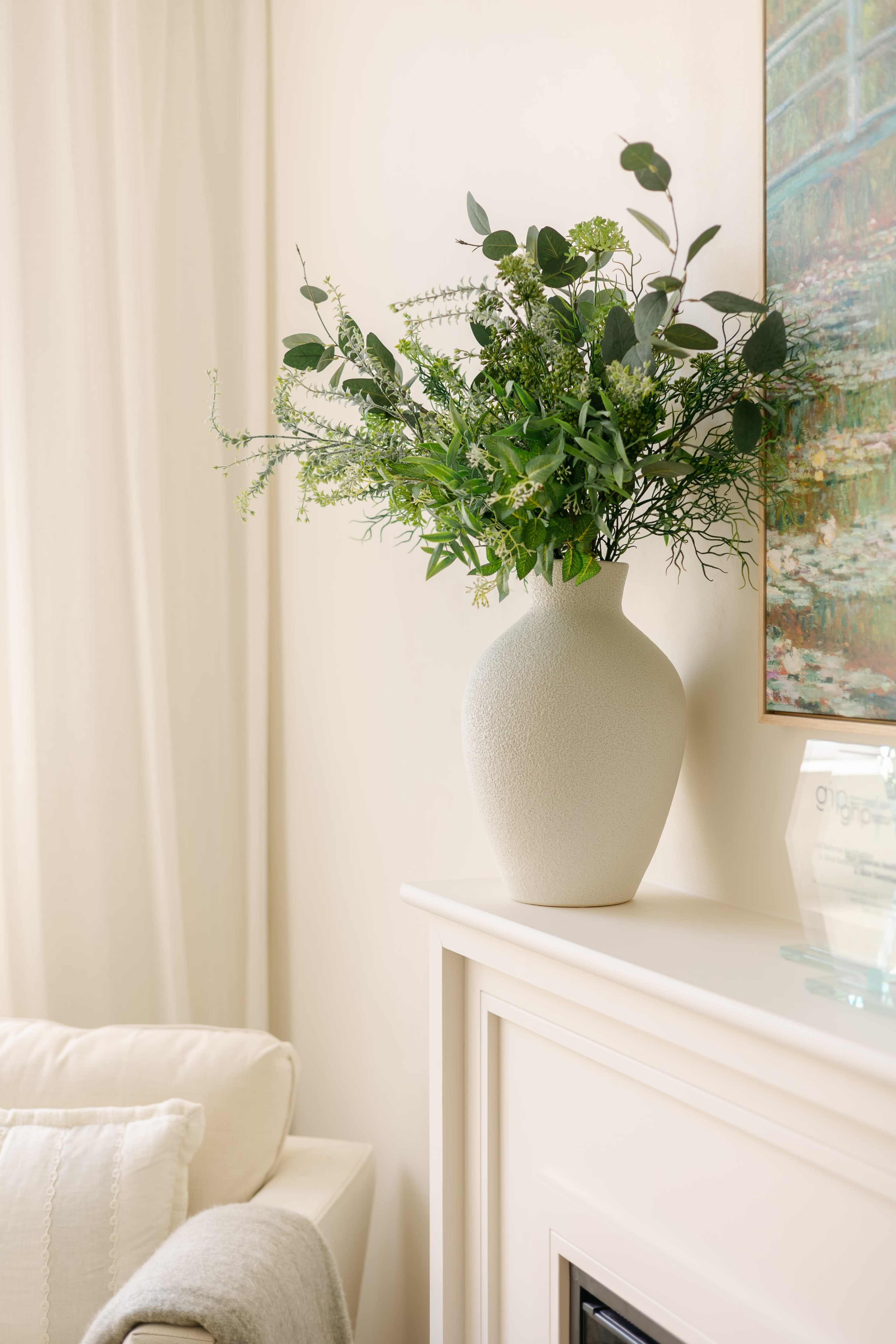 A textured white vase filled with greenery is placed on a white mantel beneath a colorful framed painting.