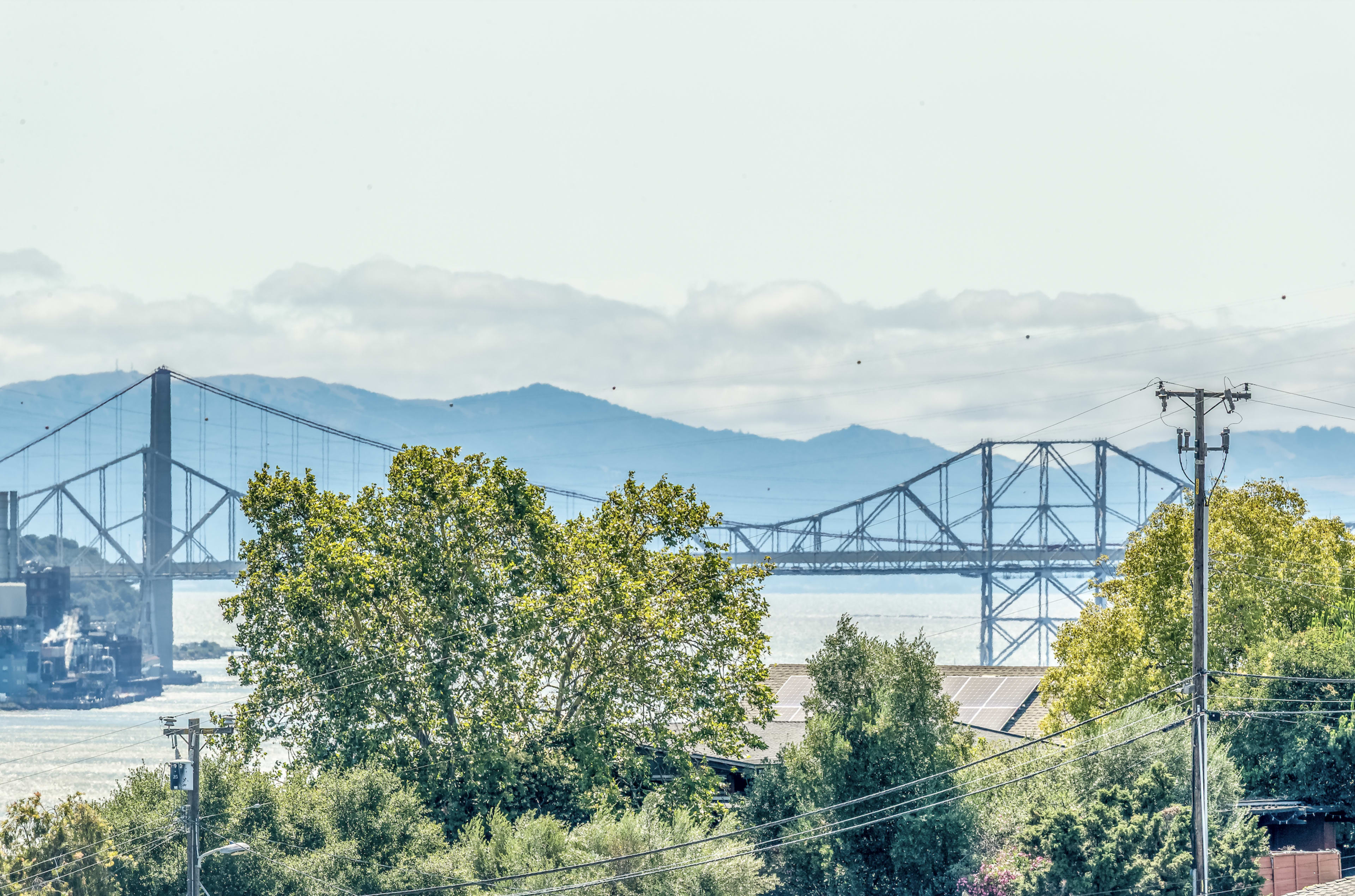 The image shows a large bridge spanning across a body of water, framed by trees and mountains in the background under a cloudy sky.