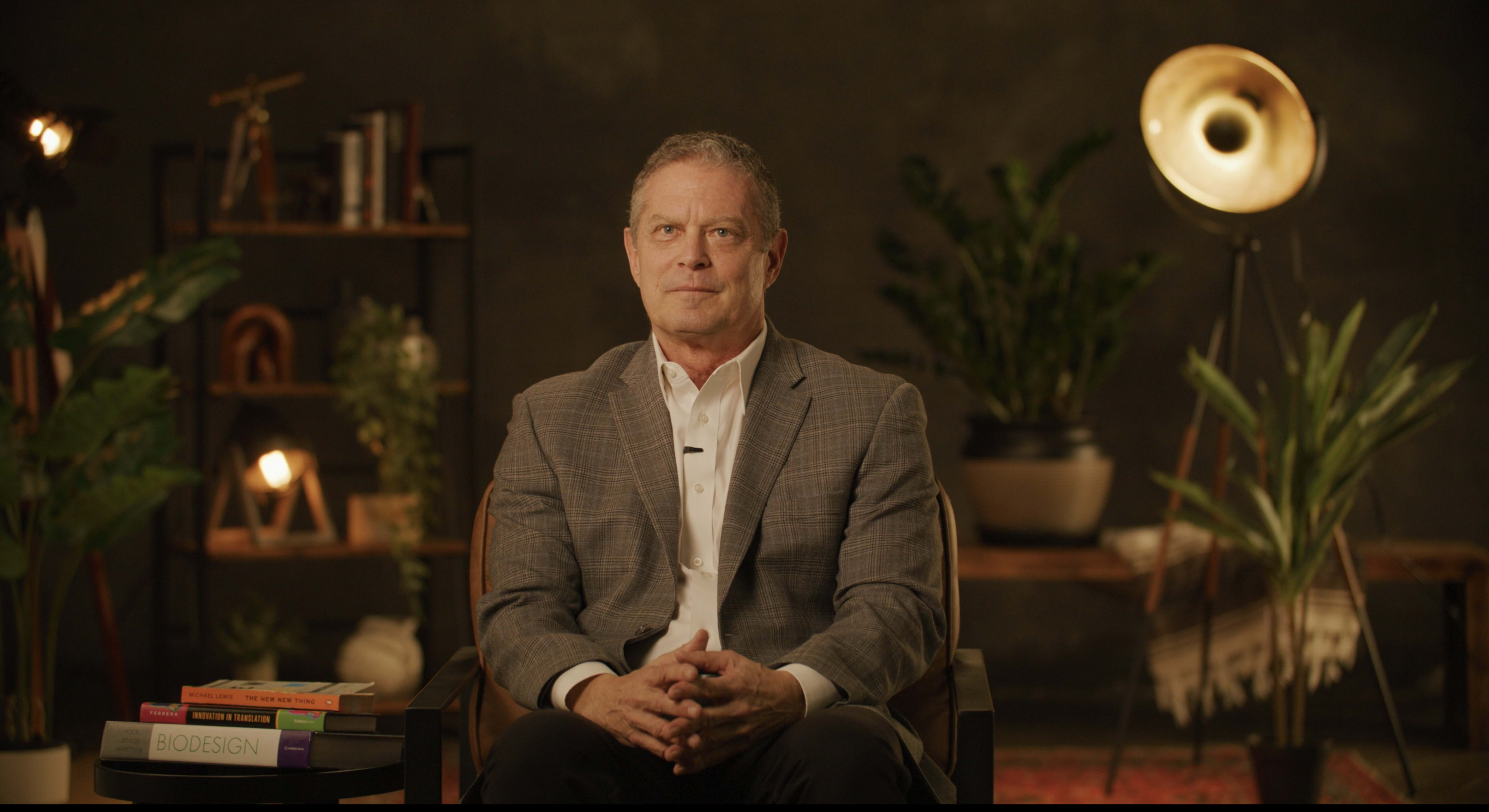 A man in a gray blazer sits confidently in a chair, surrounded by plants and books, with warm lighting in the background.