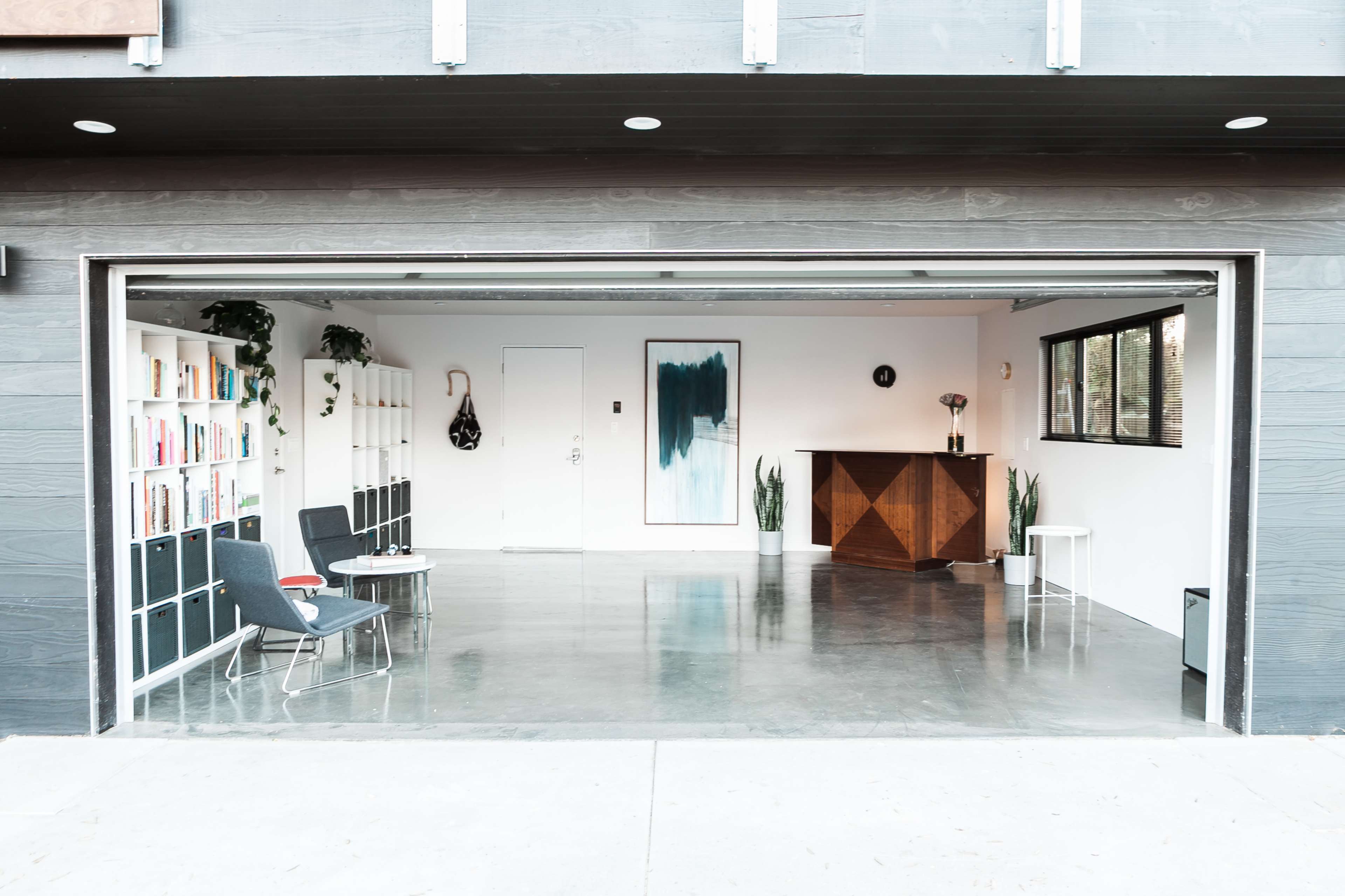 A spacious, modern indoor area with a large open garage door, featuring a bookshelf, a decorative table, and minimalist furniture.