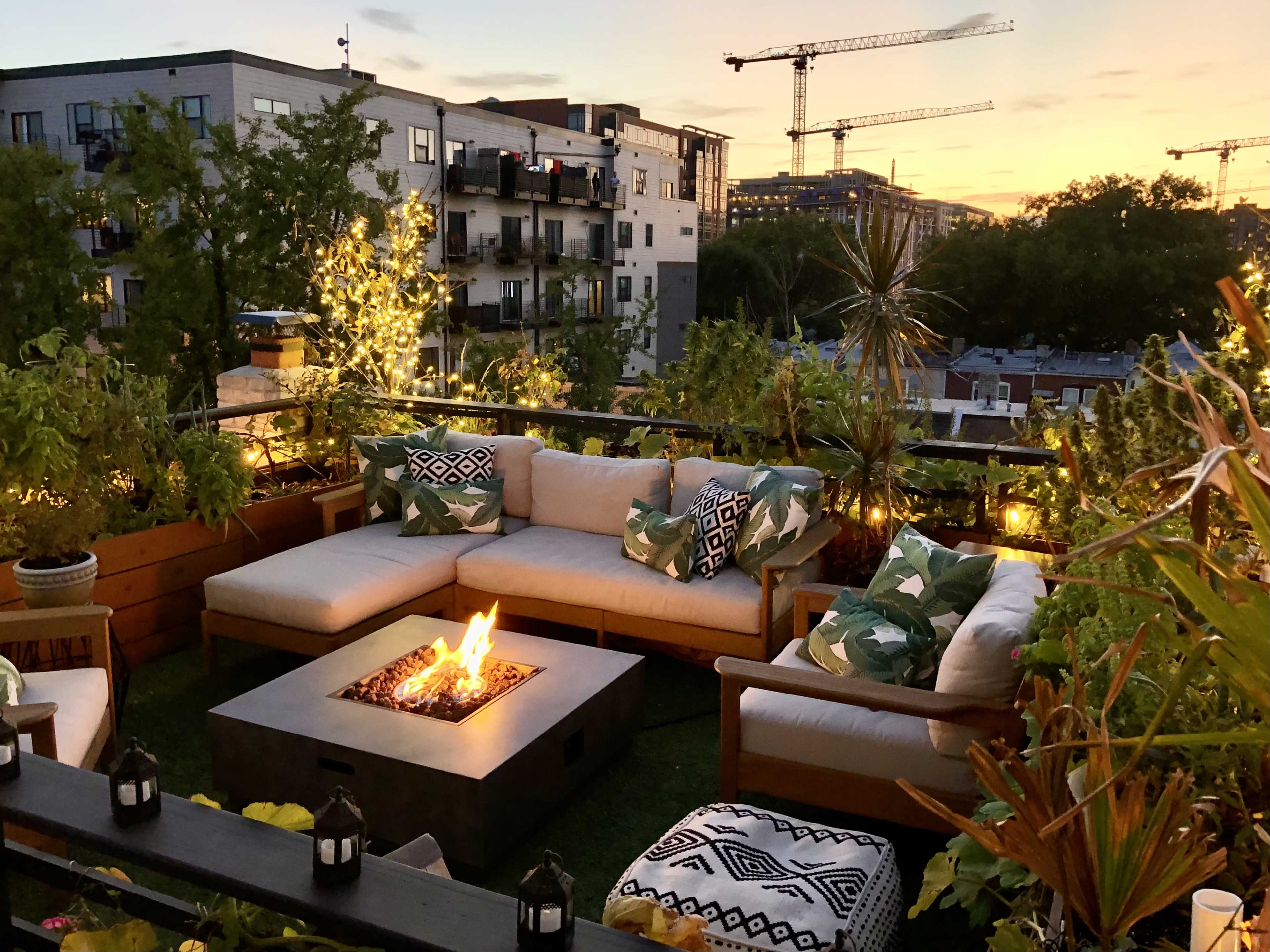 A rooftop patio with a sectional sofa, a fire pit, and potted plants, all illuminated by string lights at sunset.