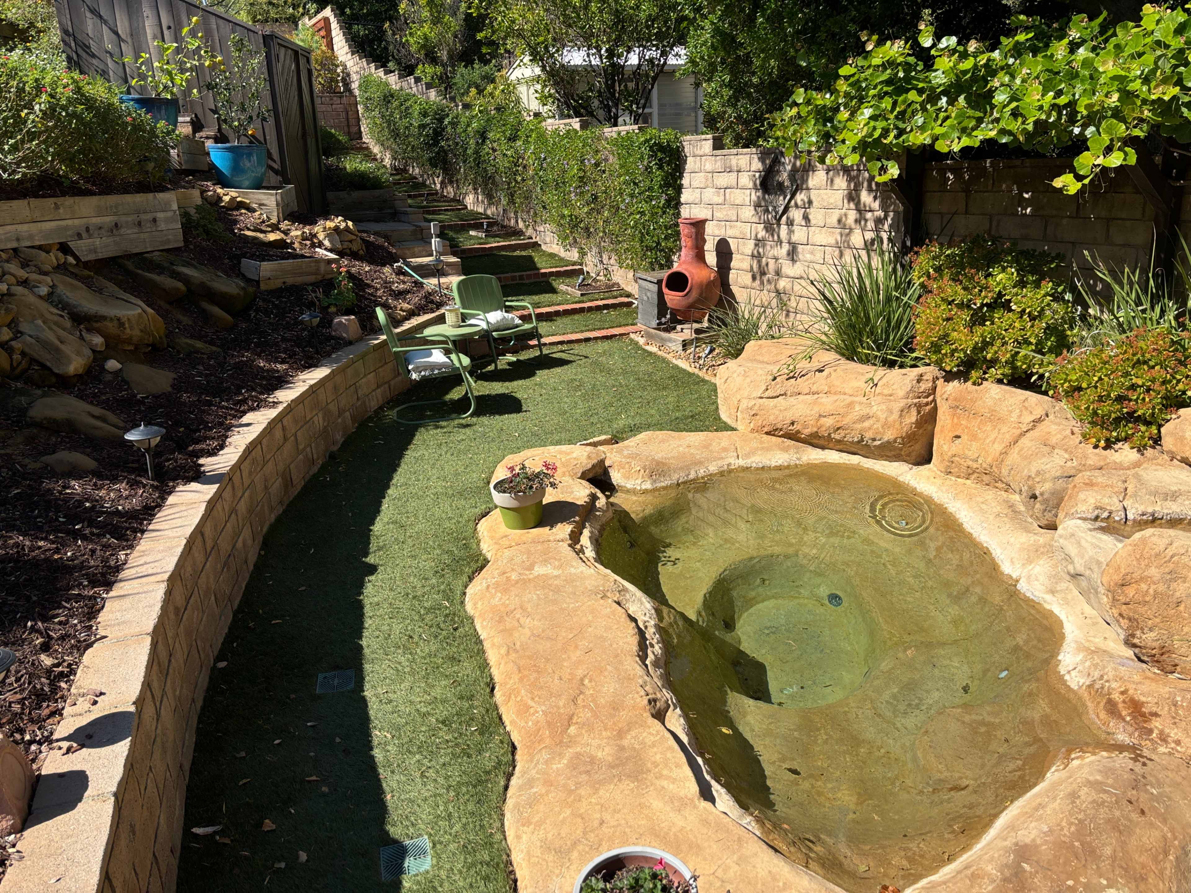 The image shows a landscaped yard with a stone-lined hot tub, a grassy path, several plants, and two green lawn chairs.