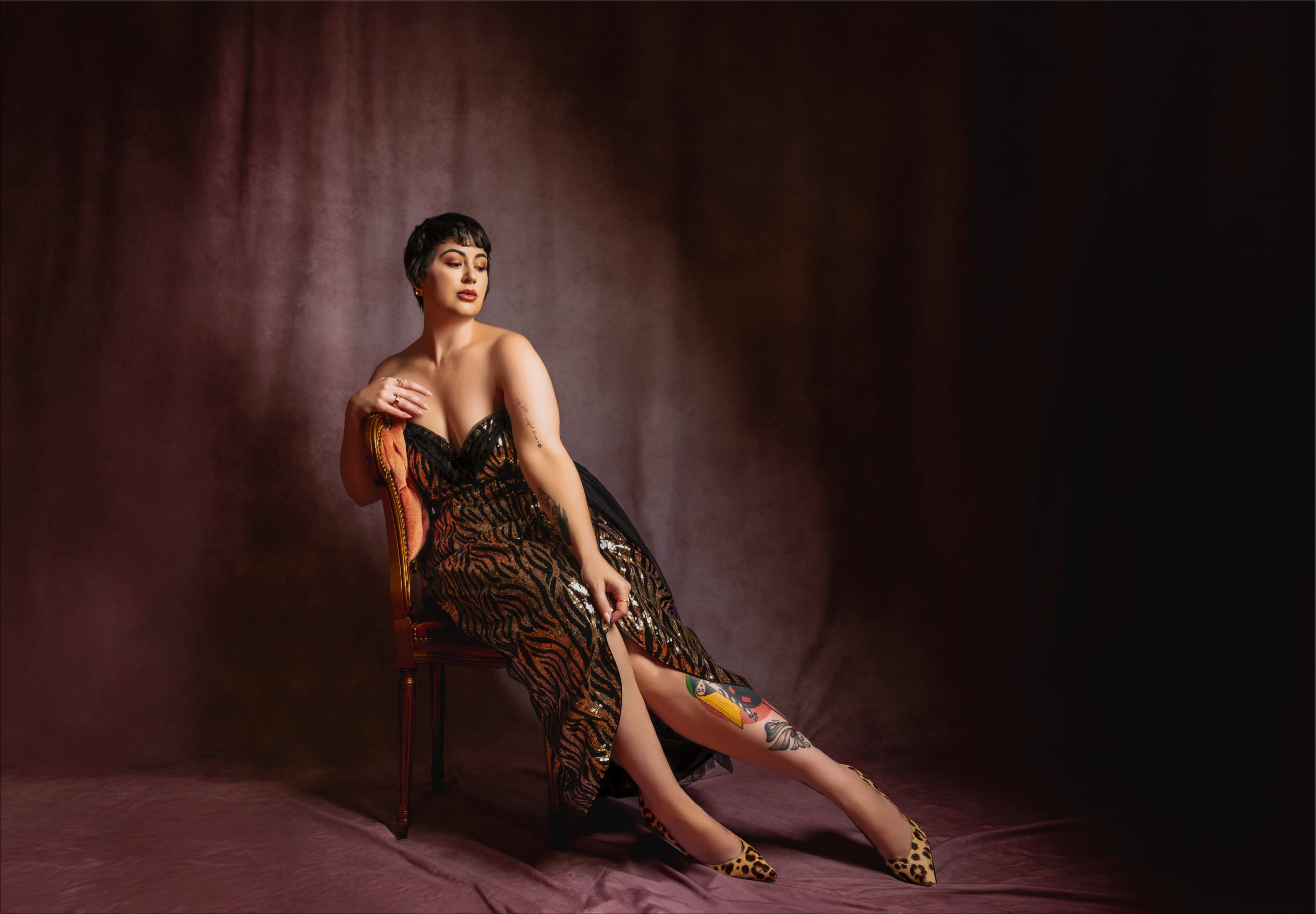 A woman in a patterned dress sits gracefully on a chair against a dark, textured backdrop.
