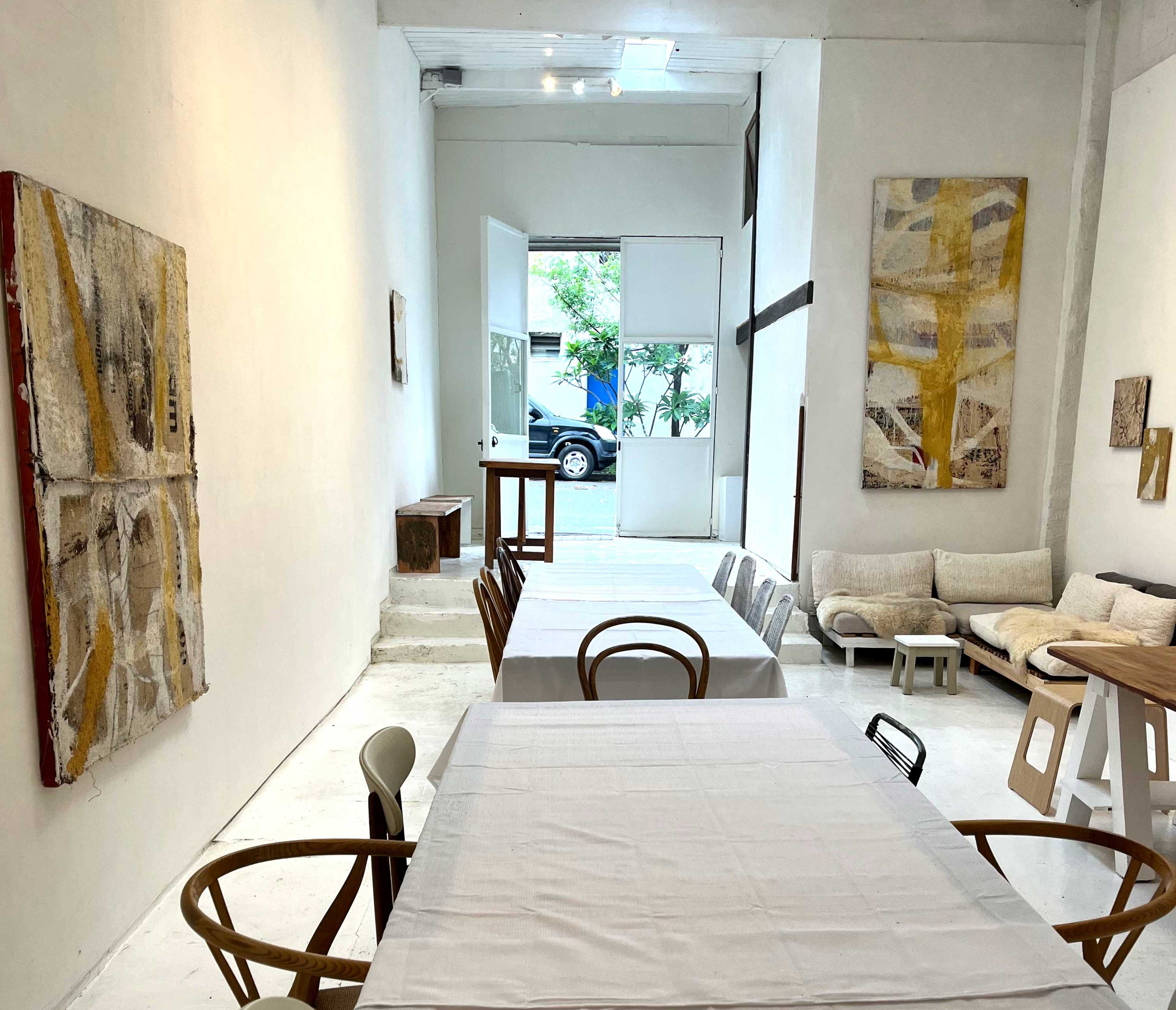 The image shows a minimalist art gallery with white walls, wooden tables set with white tablecloths, and various artworks displayed on the walls.