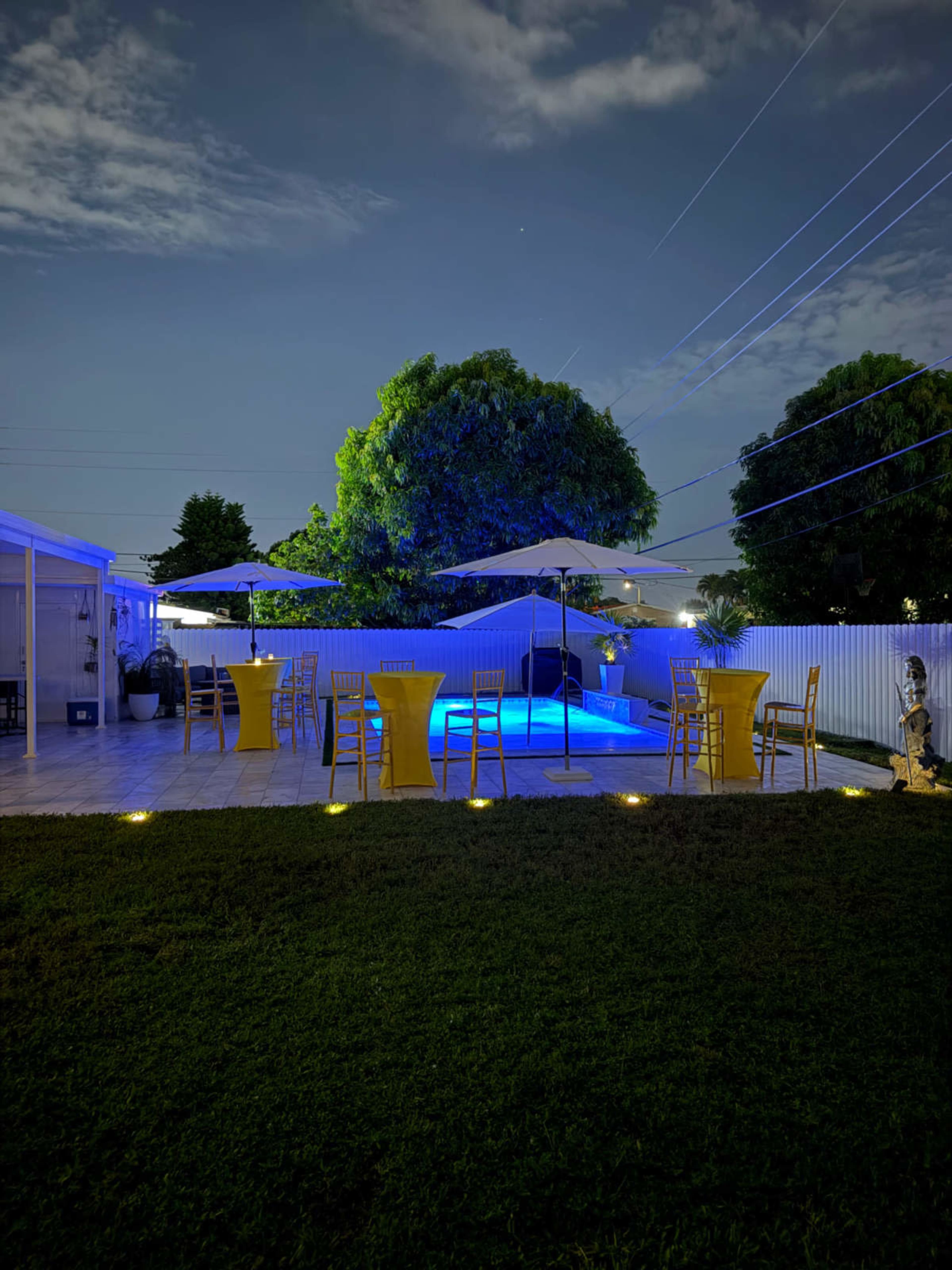 The image shows a backyard with yellow tables and chairs around a pool, illuminated by blue lighting under a night sky.