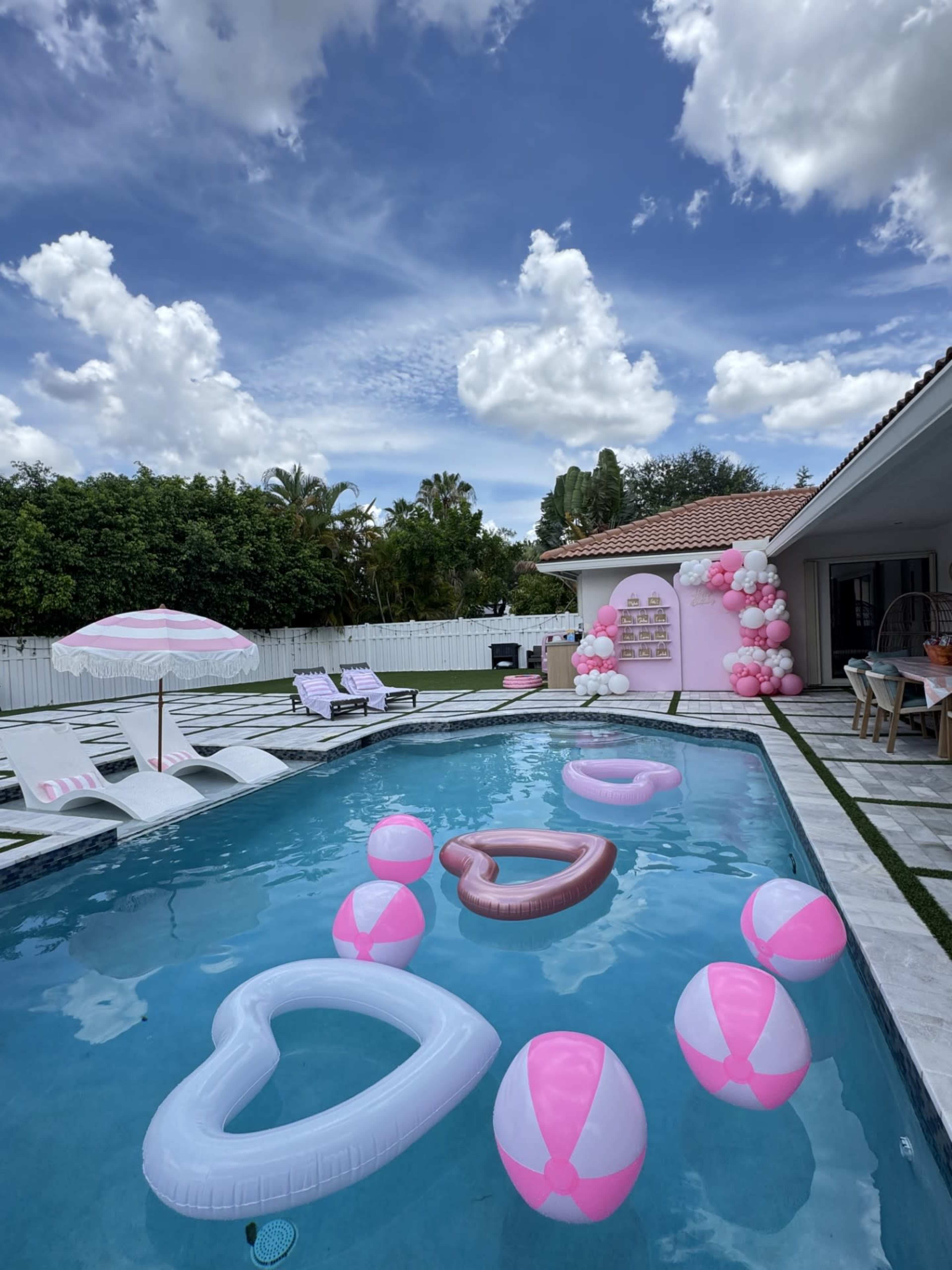 The image shows a backyard pool decorated with pink and white heart-shaped inflatables and beach balls, surrounded by lounge chairs and lush greenery.