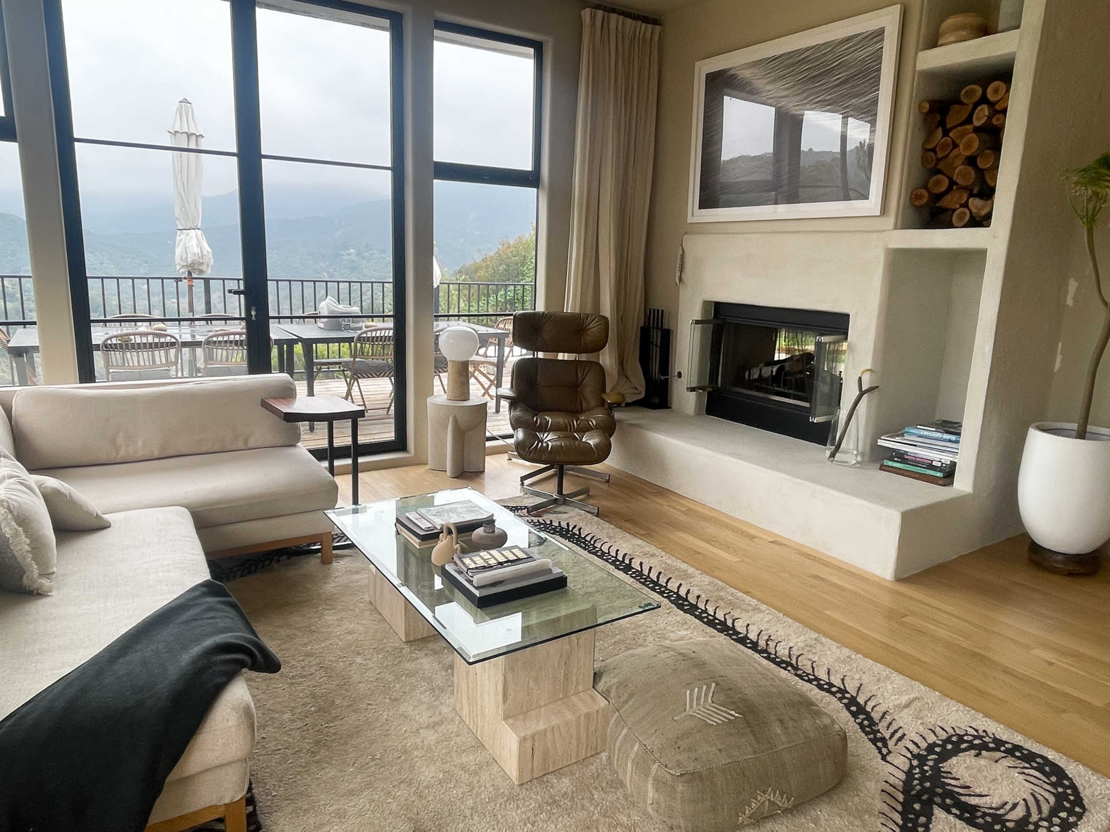 A modern living room with a glass coffee table, a beige sofa, and a fireplace, overlooking a scenic view through large windows.