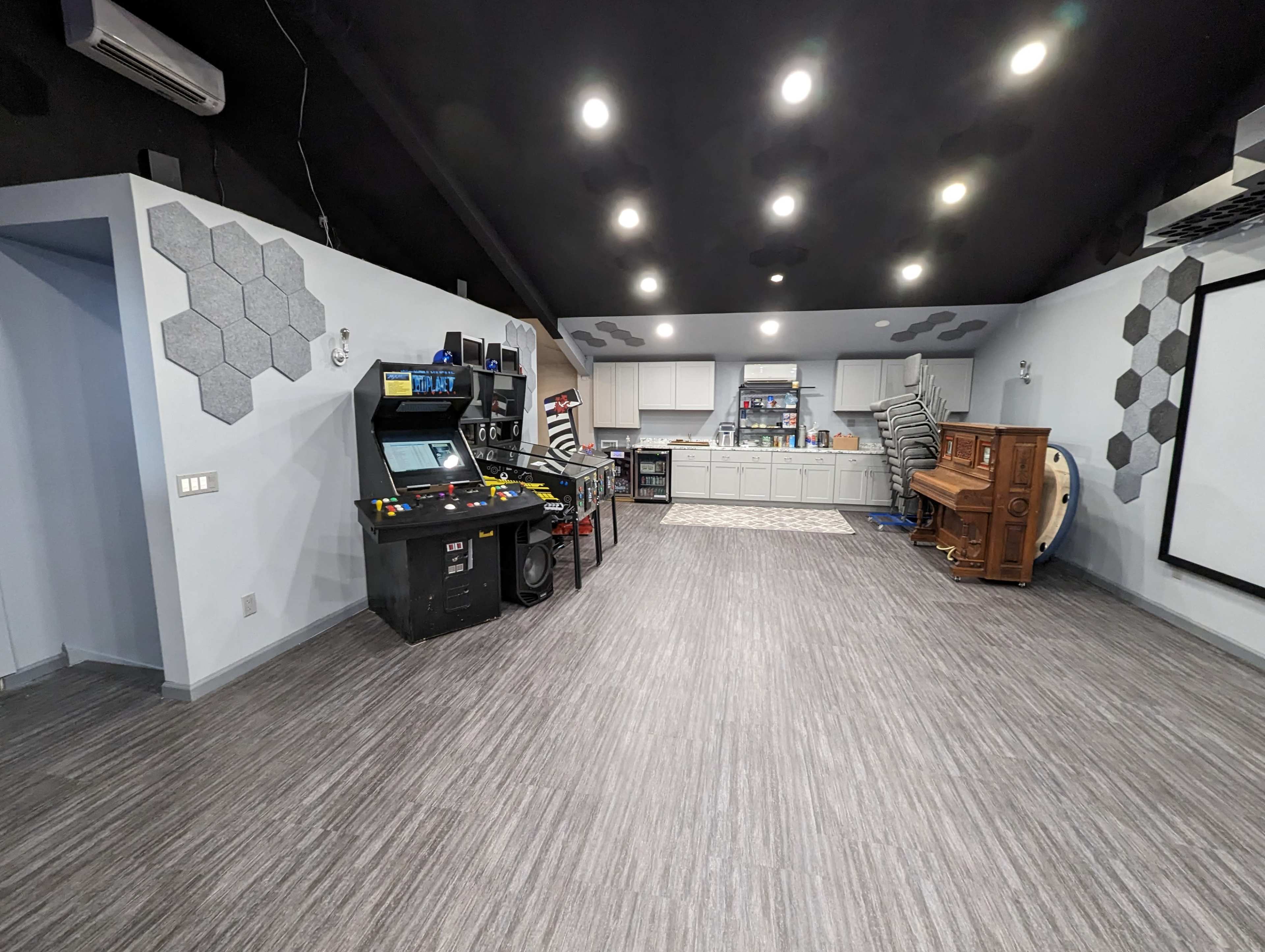 A spacious recreational room featuring a retro arcade machine, a stacked wooden piano, and a kitchenette area against a light gray wall with modern hexagonal decor.
