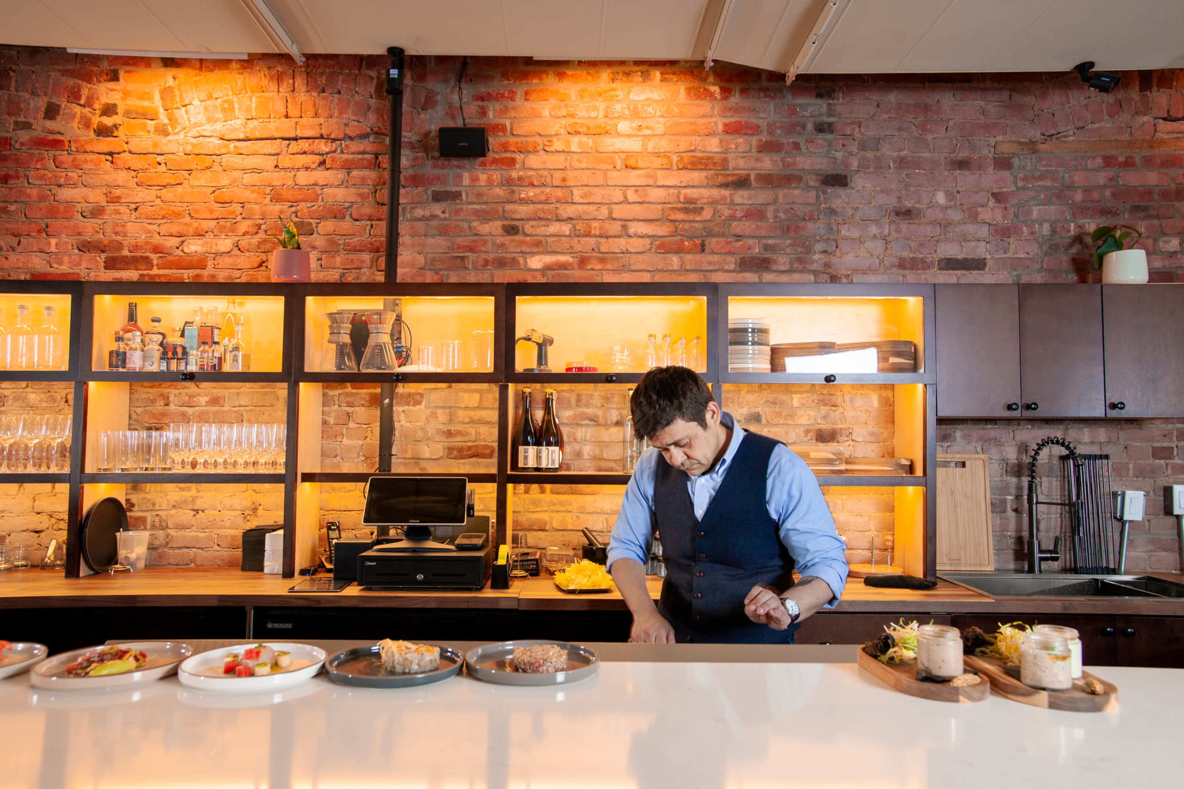 A chef prepares dishes in a kitchen with brick walls and a modern wooden display for drinks and utensils.