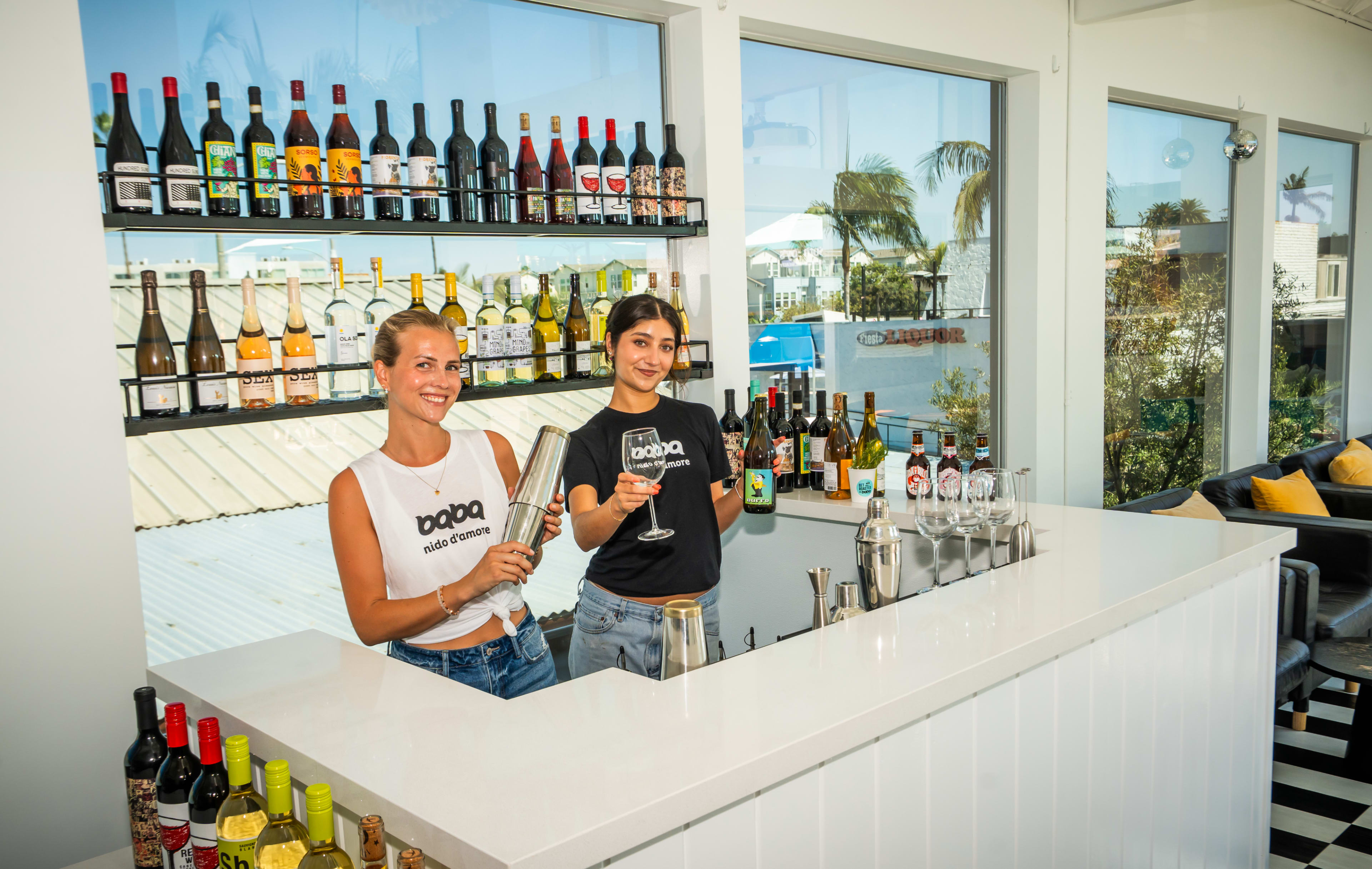 Two women stand behind a white bar with shelves of various bottles and drinks, preparing cocktails in a bright, modern space.